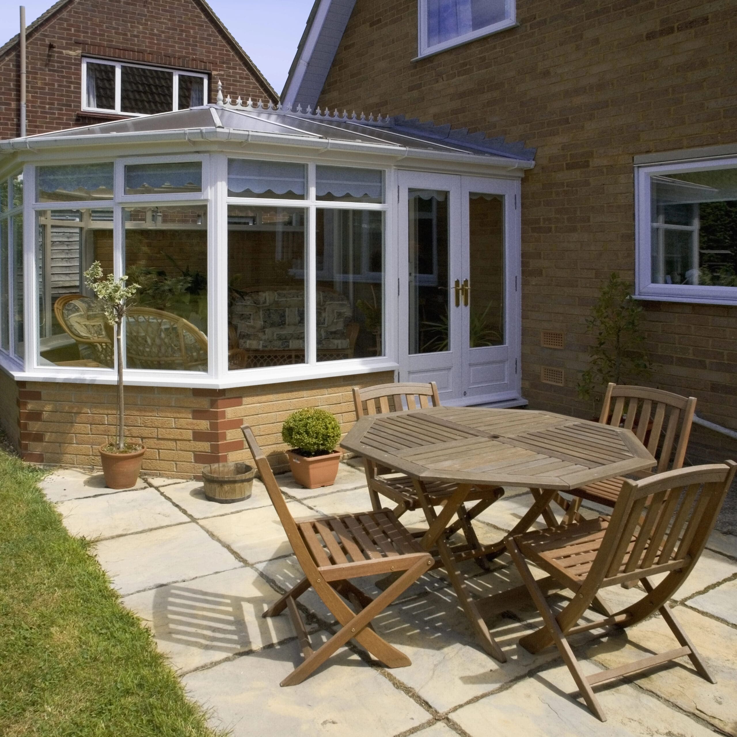 A patio with a wooden table and four chairs on a paved area next to a brick house with a glass conservatory.