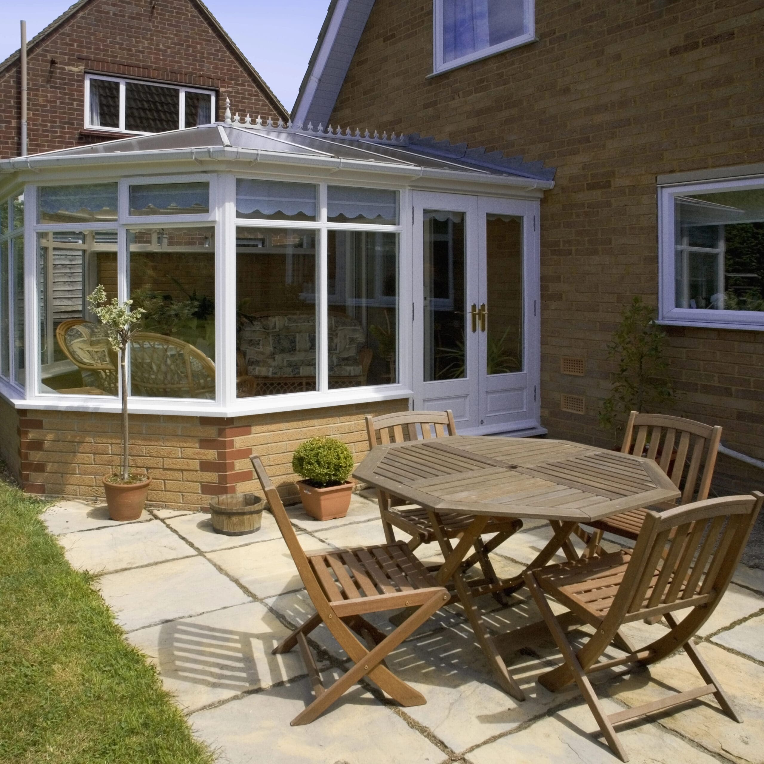 A patio with a wooden table and four chairs on a paved area next to a brick house with a glass conservatory.