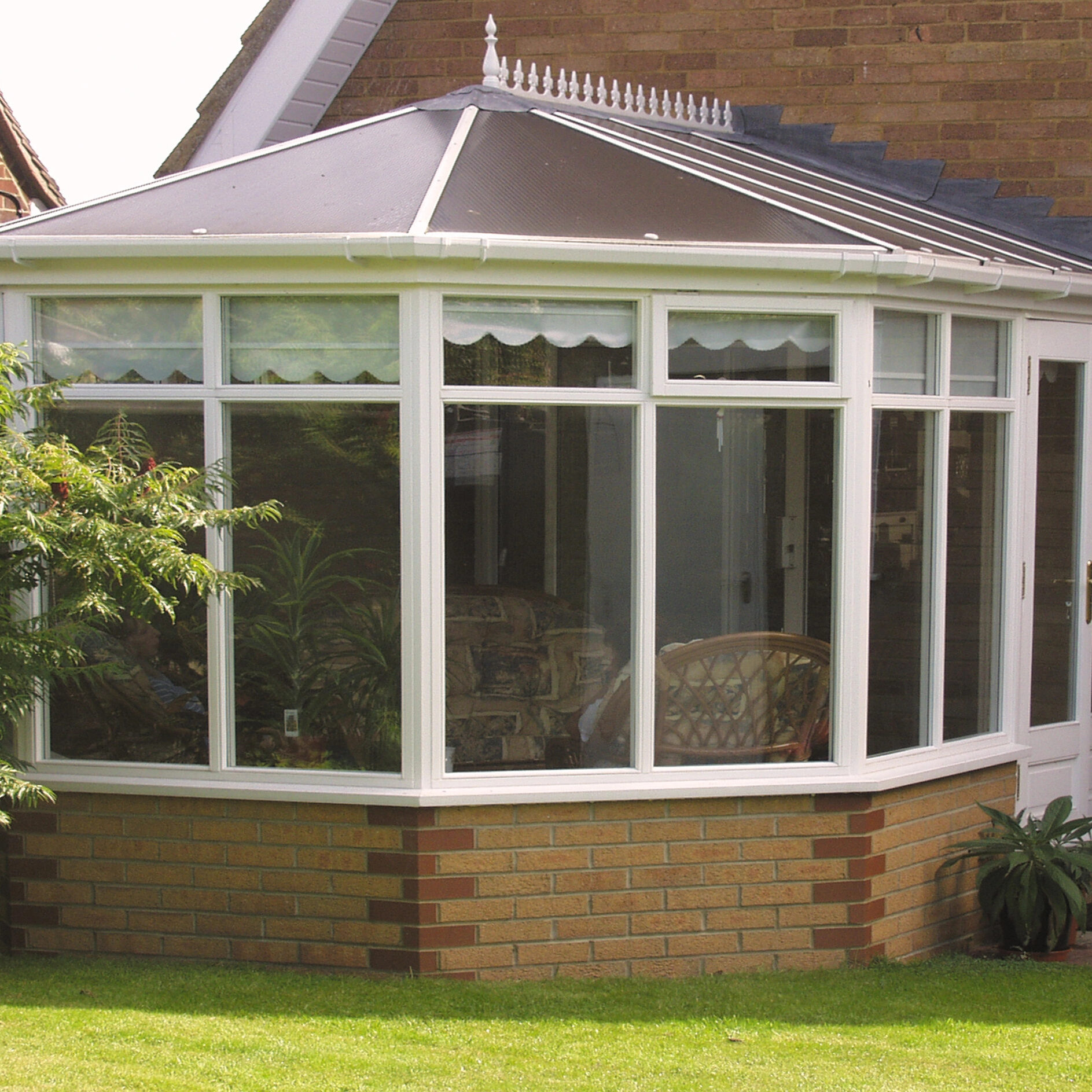 A white-framed conservatory with large windows extends from a brick house. The space includes seating and plants inside, with a small tree and green lawn in the foreground.