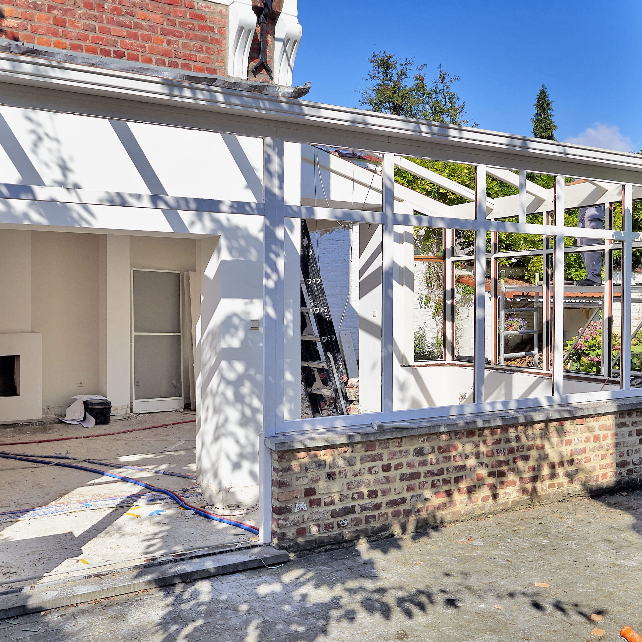 Partially constructed sunroom with large windows and white frames attached to a brick house, surrounded by garden greenery under a clear sky.