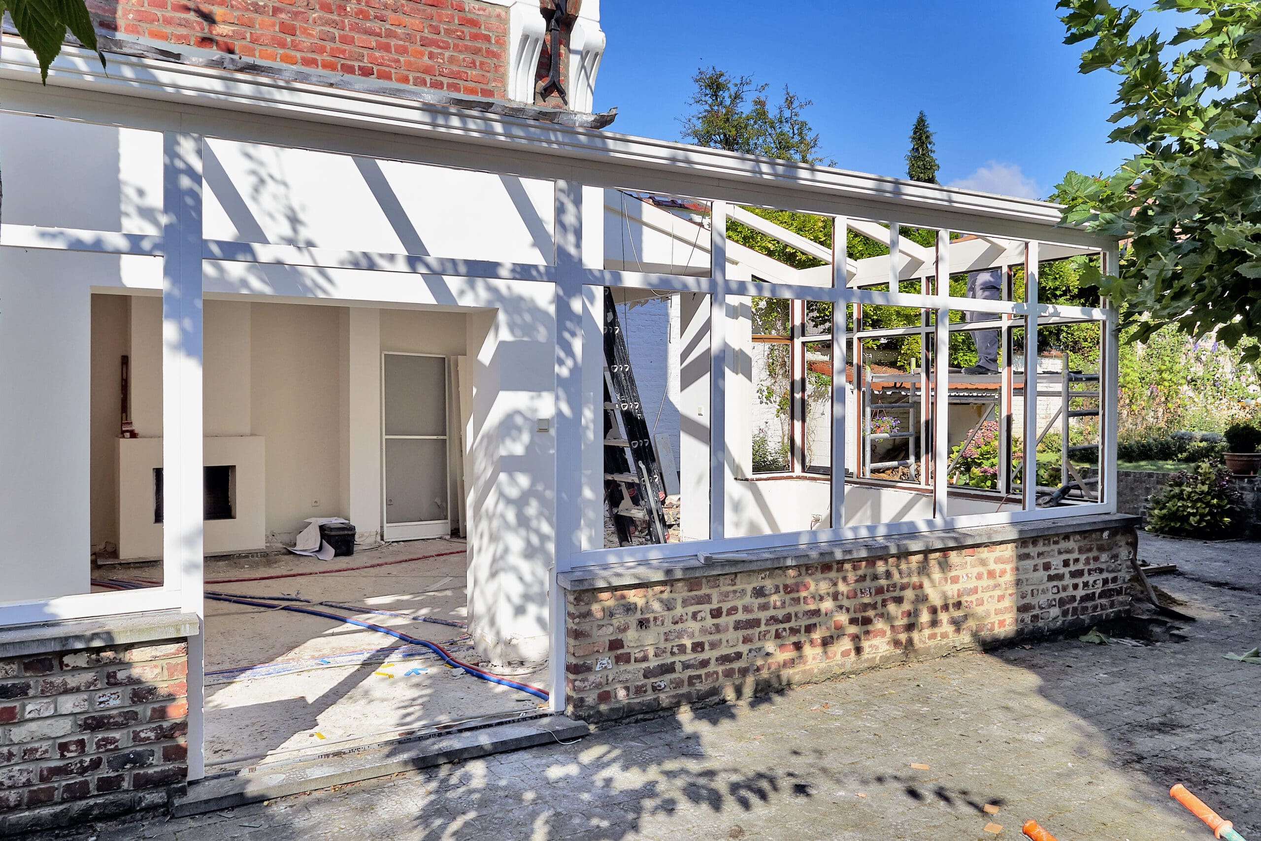 Partially constructed sunroom with large windows and white frames attached to a brick house, surrounded by garden greenery under a clear sky.