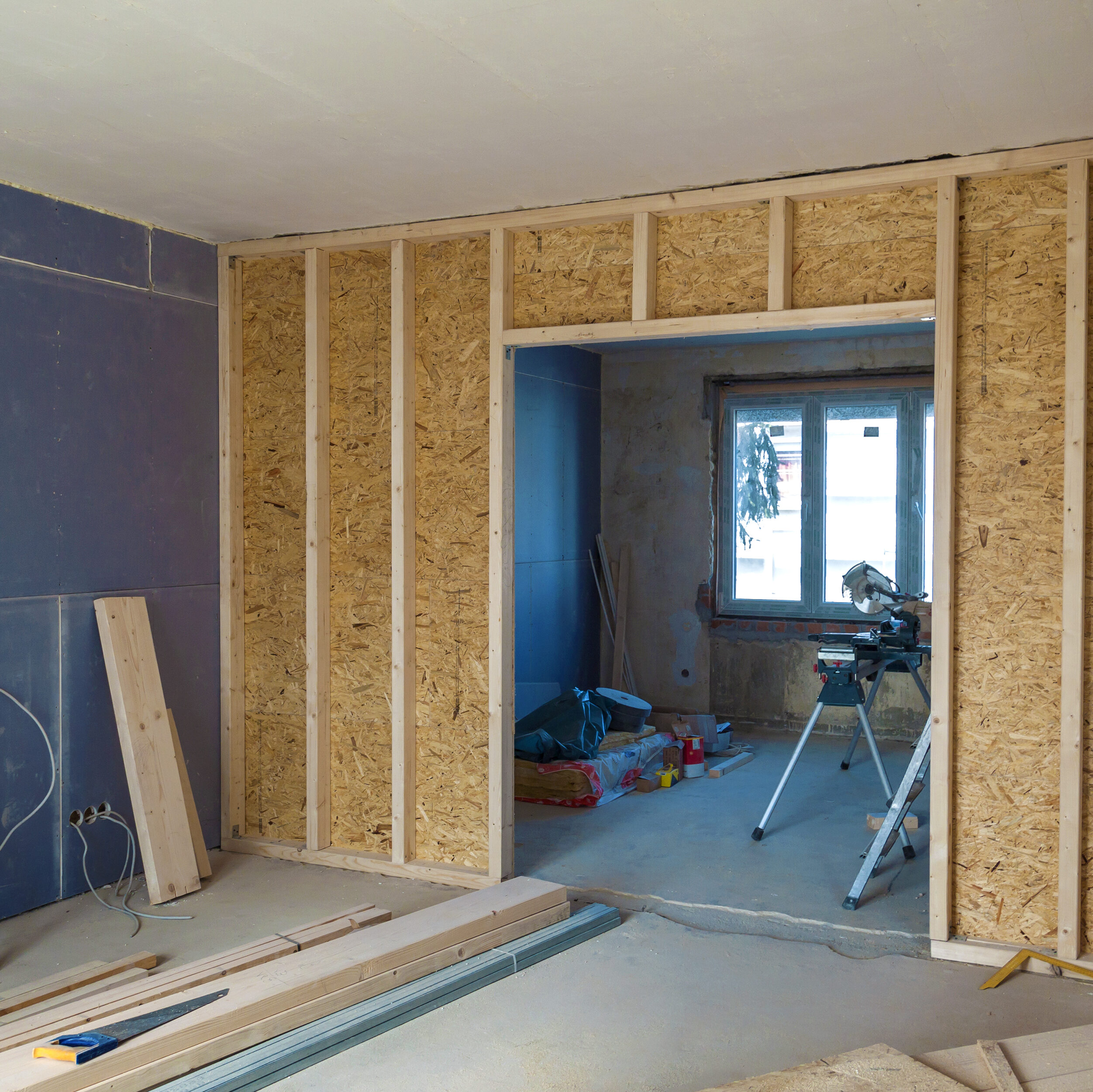 Interior view of a room under construction with framed walls, exposed insulation, and scattered tools. An open doorway leads to another unfinished room with a window.