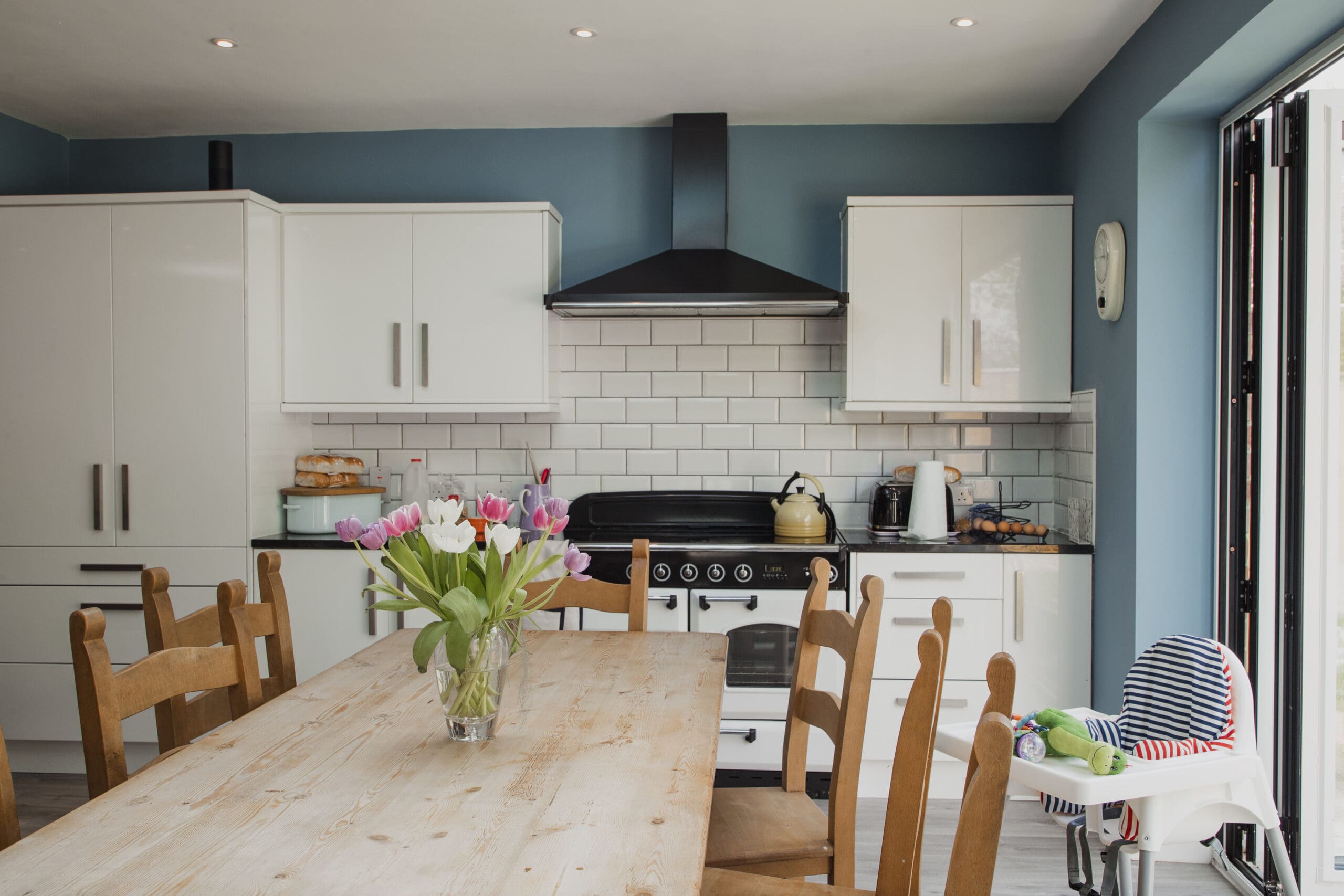 A modern kitchen with white cabinets, a black range hood, a vase of flowers on a wooden dining table, and a high chair on one side. The walls are painted blue, and there are ceiling lights.