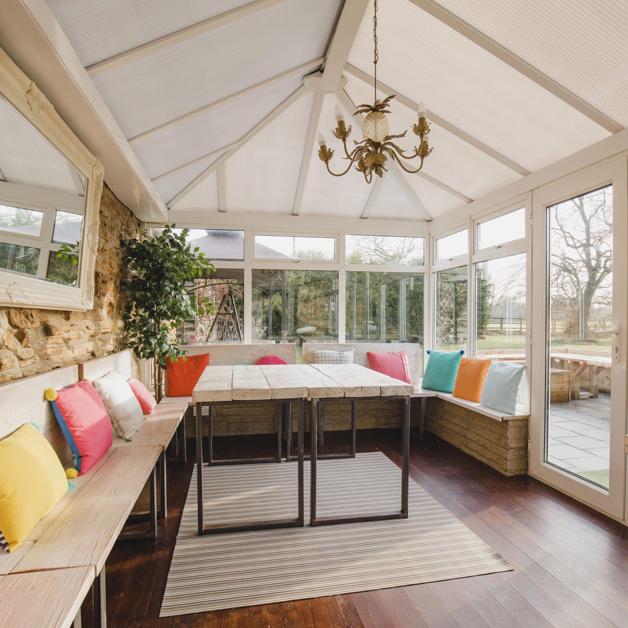 A sunlit conservatory with a rustic table and benches adorned with colorful cushions, surrounded by large windows and a hanging chandelier.