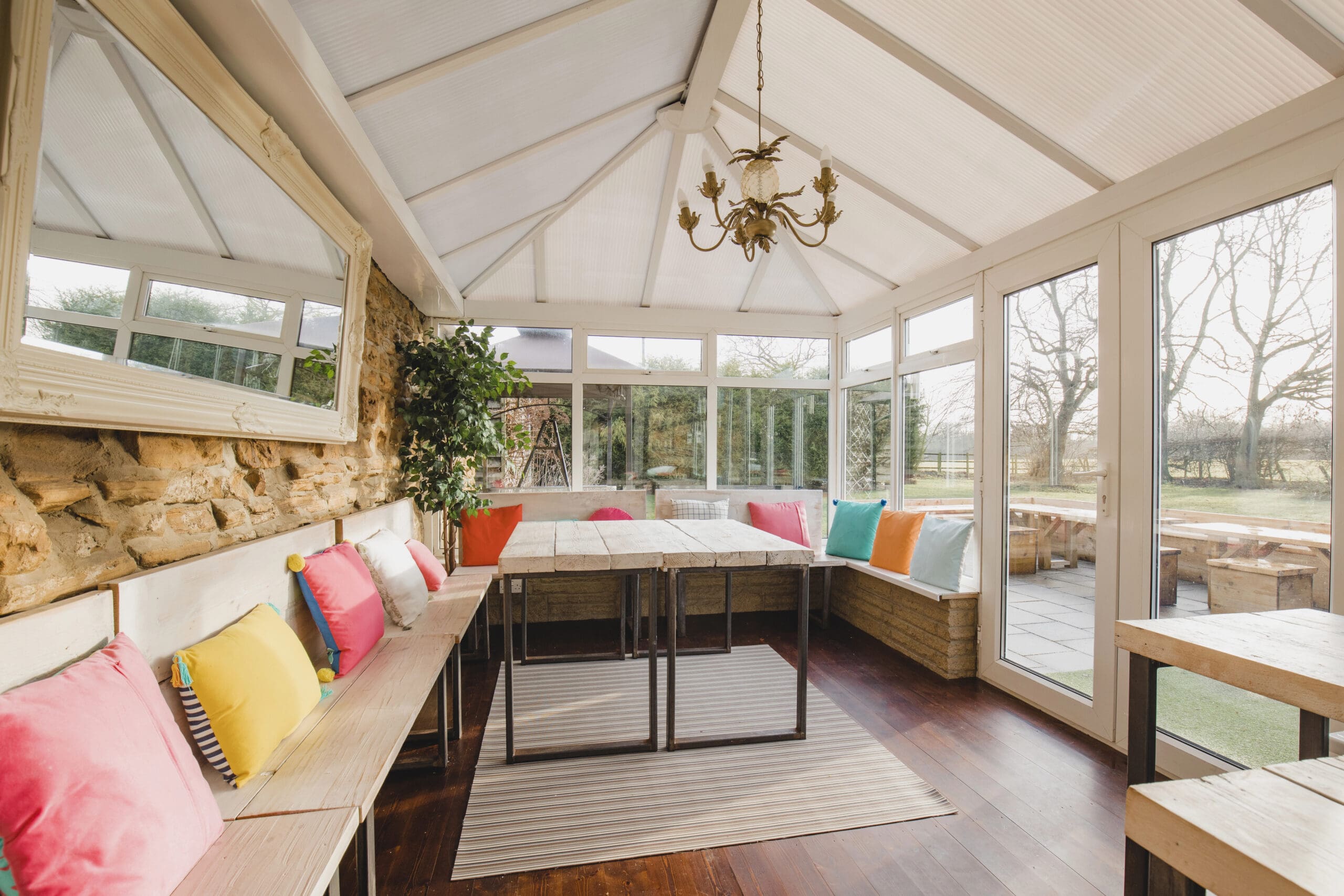 A sunlit conservatory with a rustic table and benches adorned with colorful cushions, surrounded by large windows and a hanging chandelier.