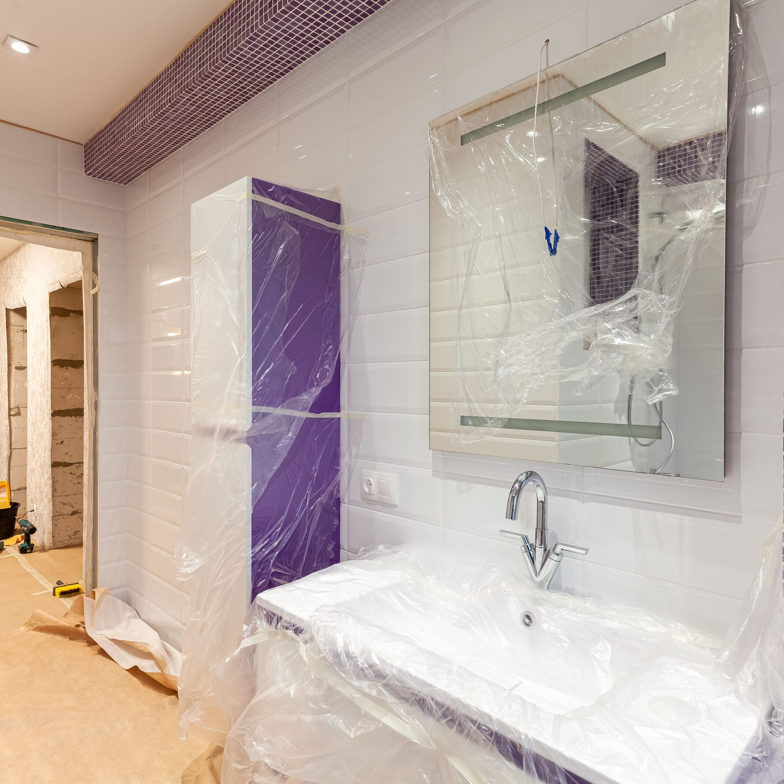 A bathroom under renovation with plastic sheets covering cabinets and a mirror. A sink with a faucet is installed, and construction materials are visible in the background.