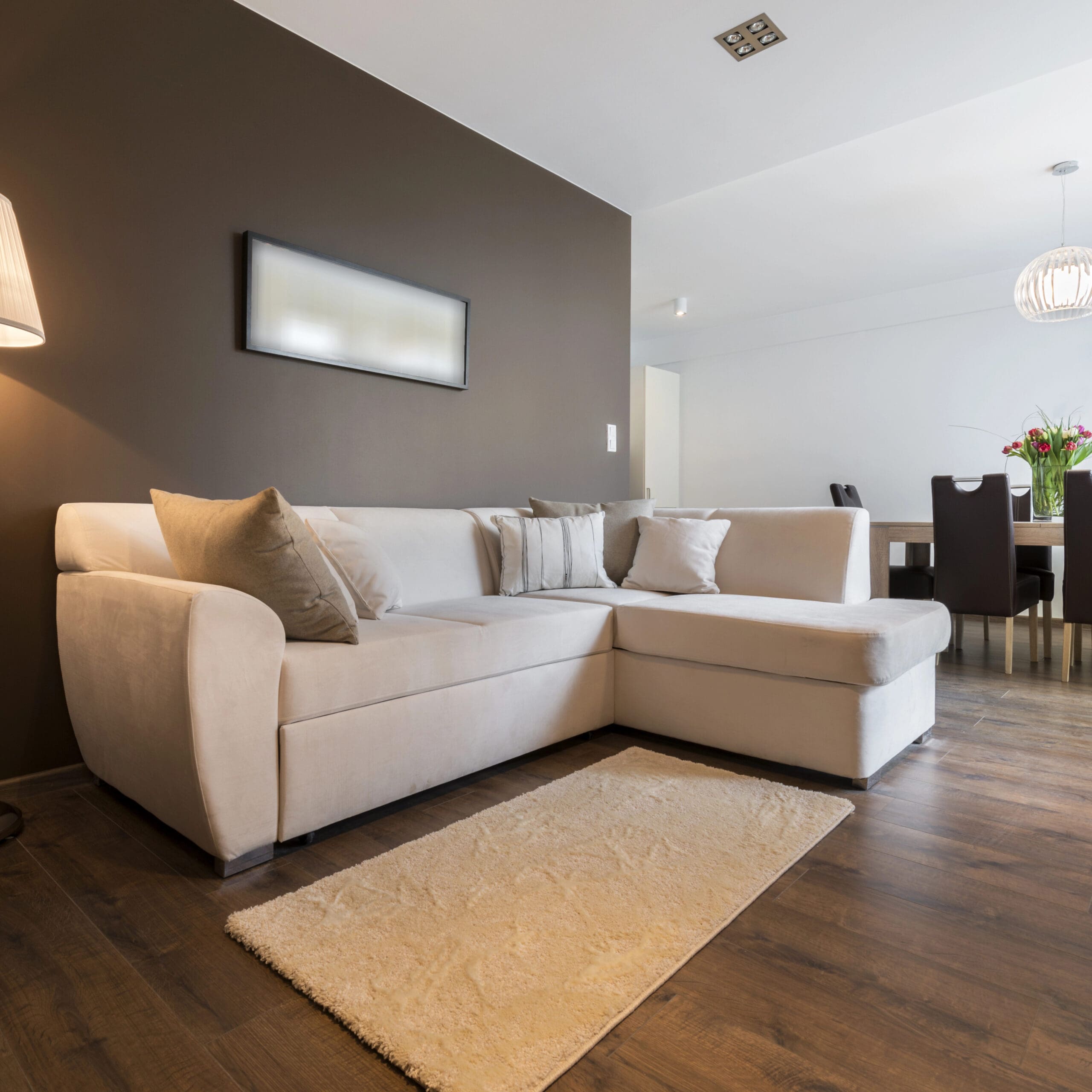 A modern living room features a beige sectional sofa with throw pillows, a small rug, a brown accent wall, and wooden flooring. A dining area with a table and chairs is visible in the background.