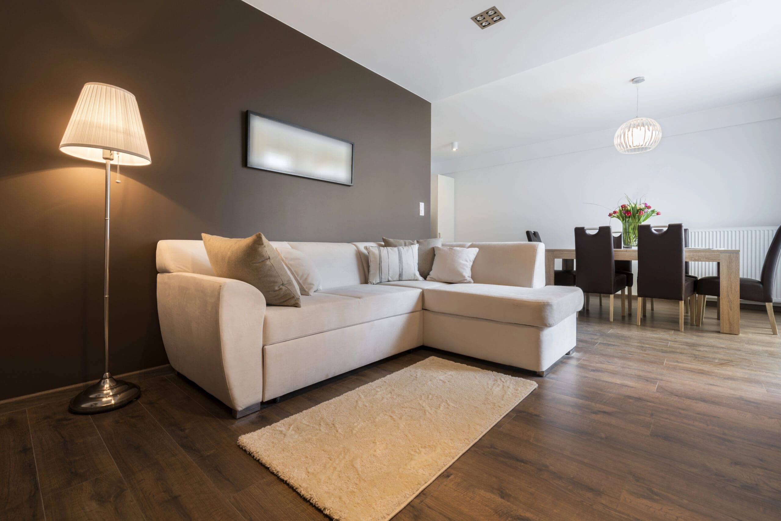 A modern living area with a beige sectional sofa, a floor lamp, a small rug on wooden flooring, and a dining area in the background with a light fixture and a table with six chairs.