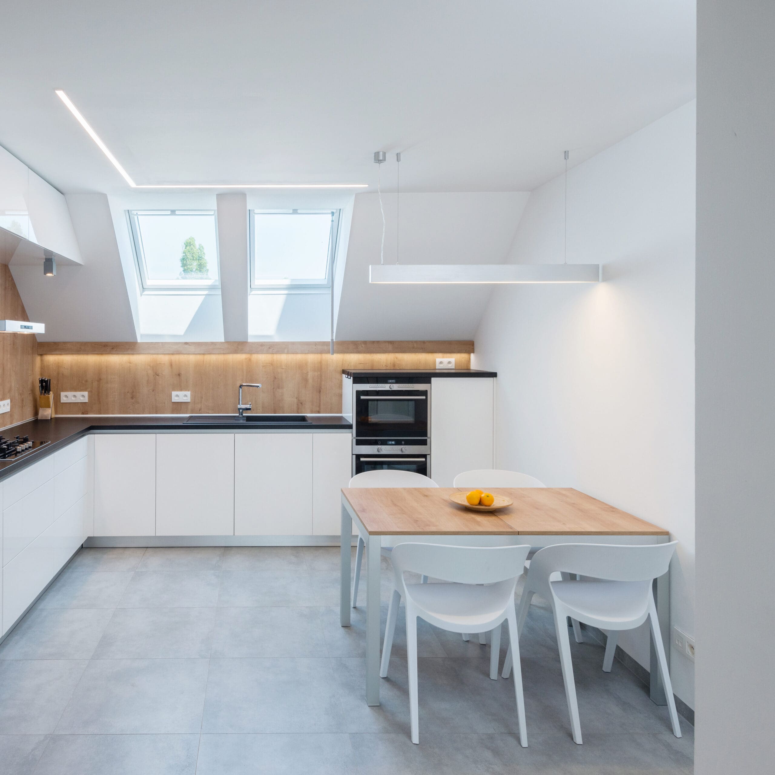 A modern kitchen with white cabinets, a wooden countertop, a built-in oven, skylight windows, and a small wooden dining table with four white chairs. A white pendant light hangs above the table.