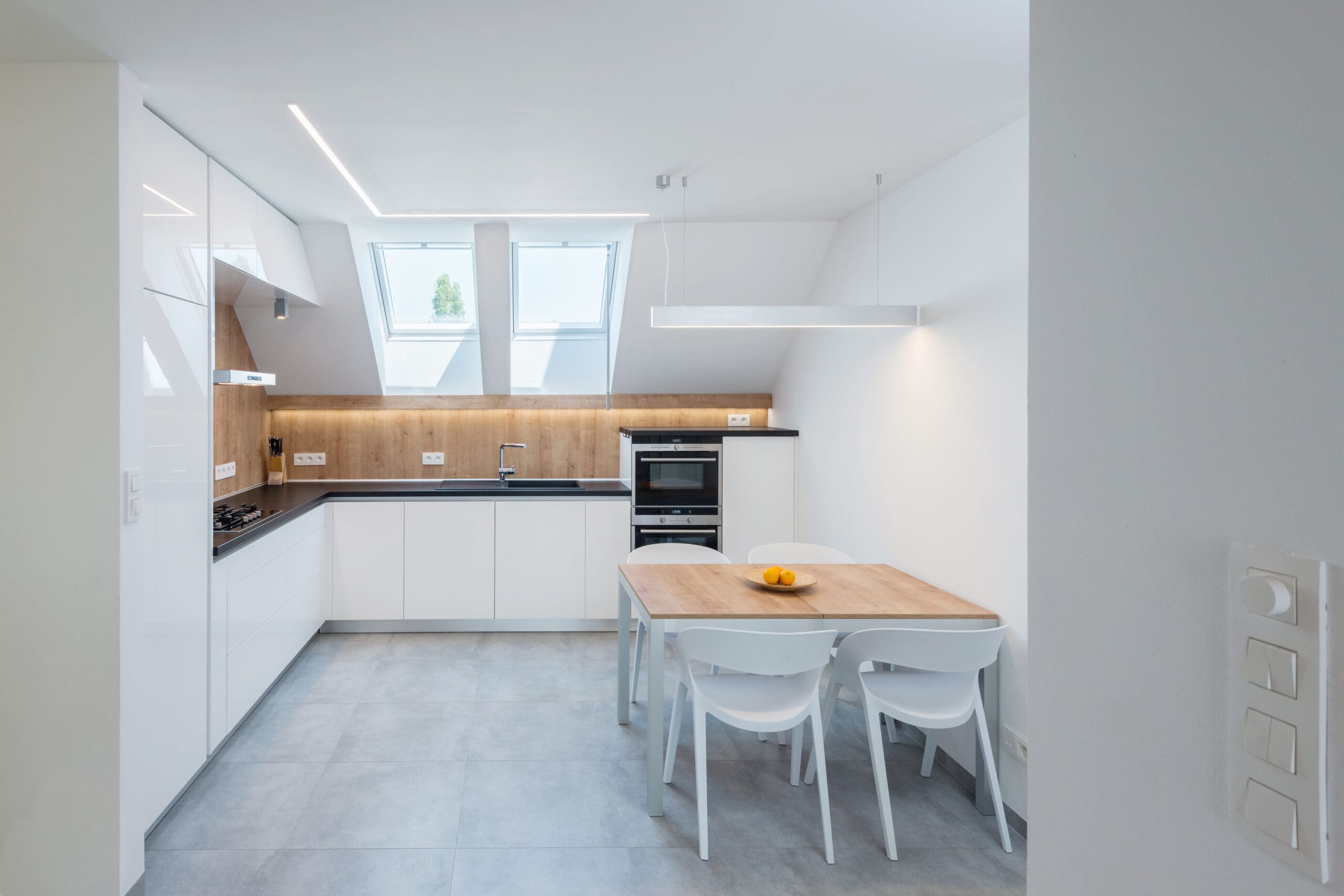 Modern kitchen with white cabinets, an L-shaped countertop, an oven, and a sink under skylights. There is a wooden dining table with four white chairs and a bowl of fruit.