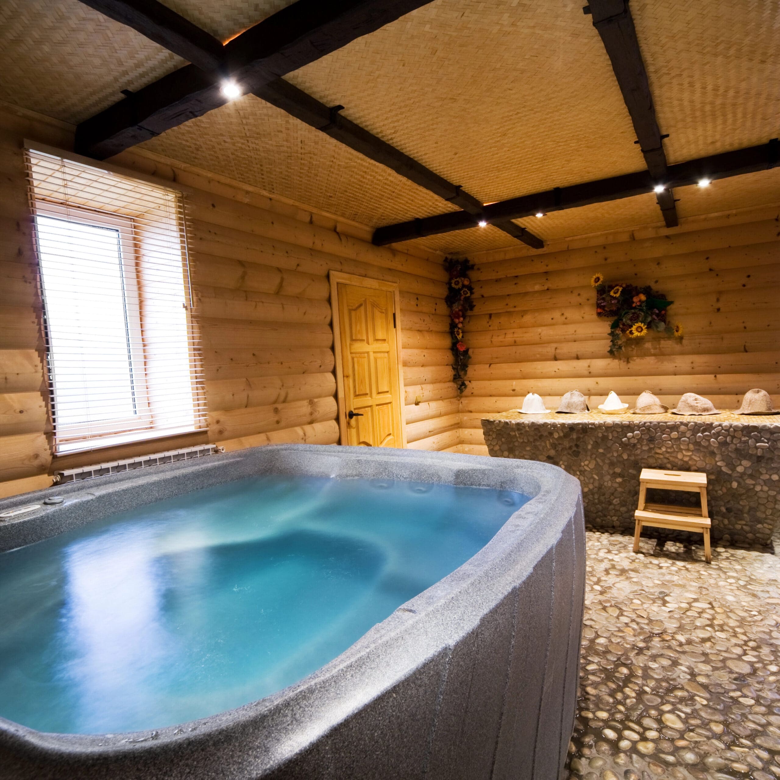 A cozy wooden room with a hot tub, stone flooring, and two windows with blinds. A bench sits in front of a decorative table lined with candles. A door is centered on the far wall.