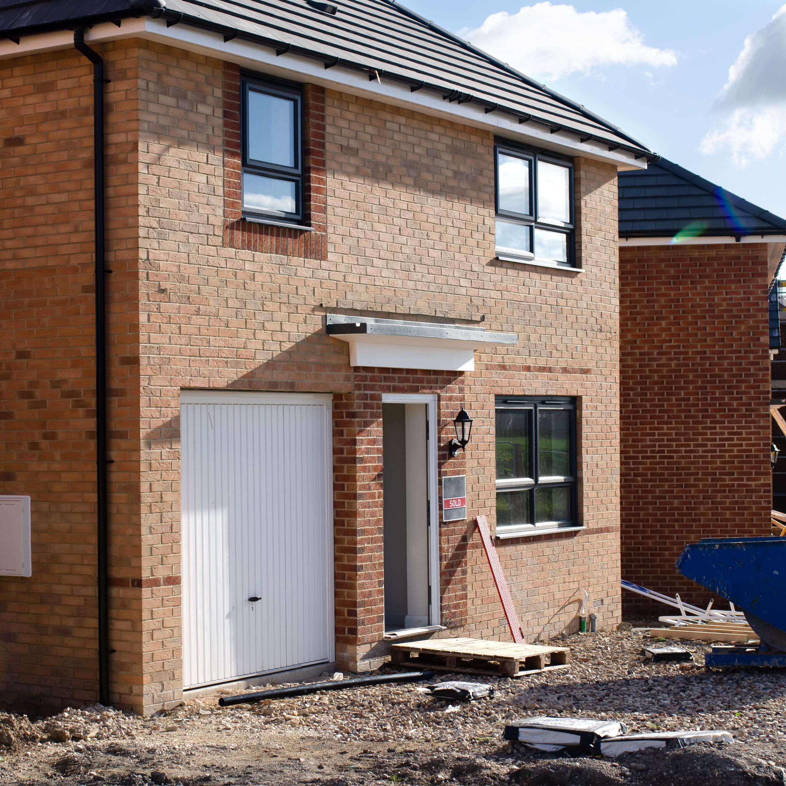 A newly constructed brick house with a garage door, front door without steps, and unfinished yard. Construction equipment and materials are visible nearby.