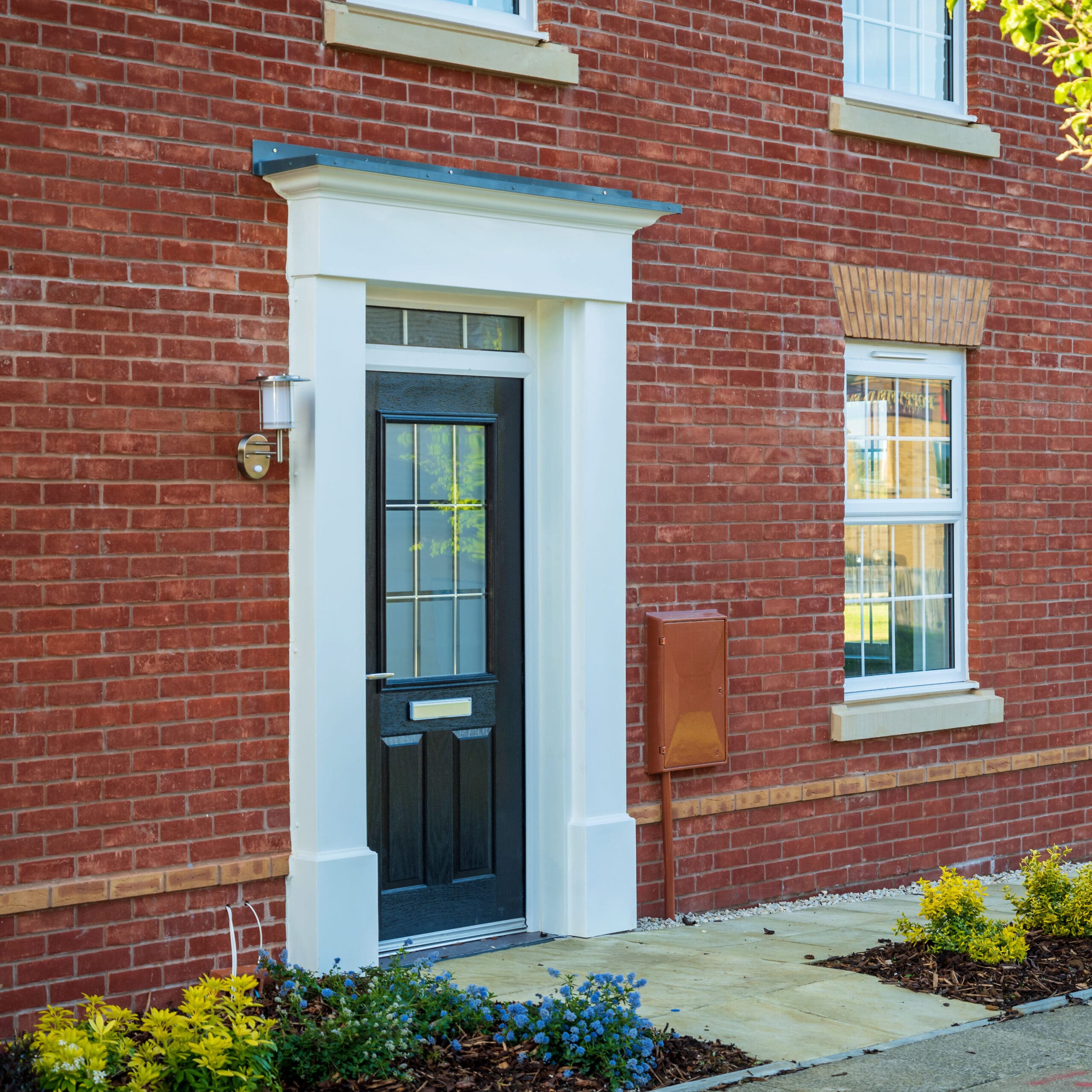 Front view of a brick house with a black front door framed in white, a nearby window with white trim, outdoor light fixture, mailbox, and small garden with yellow flowers.