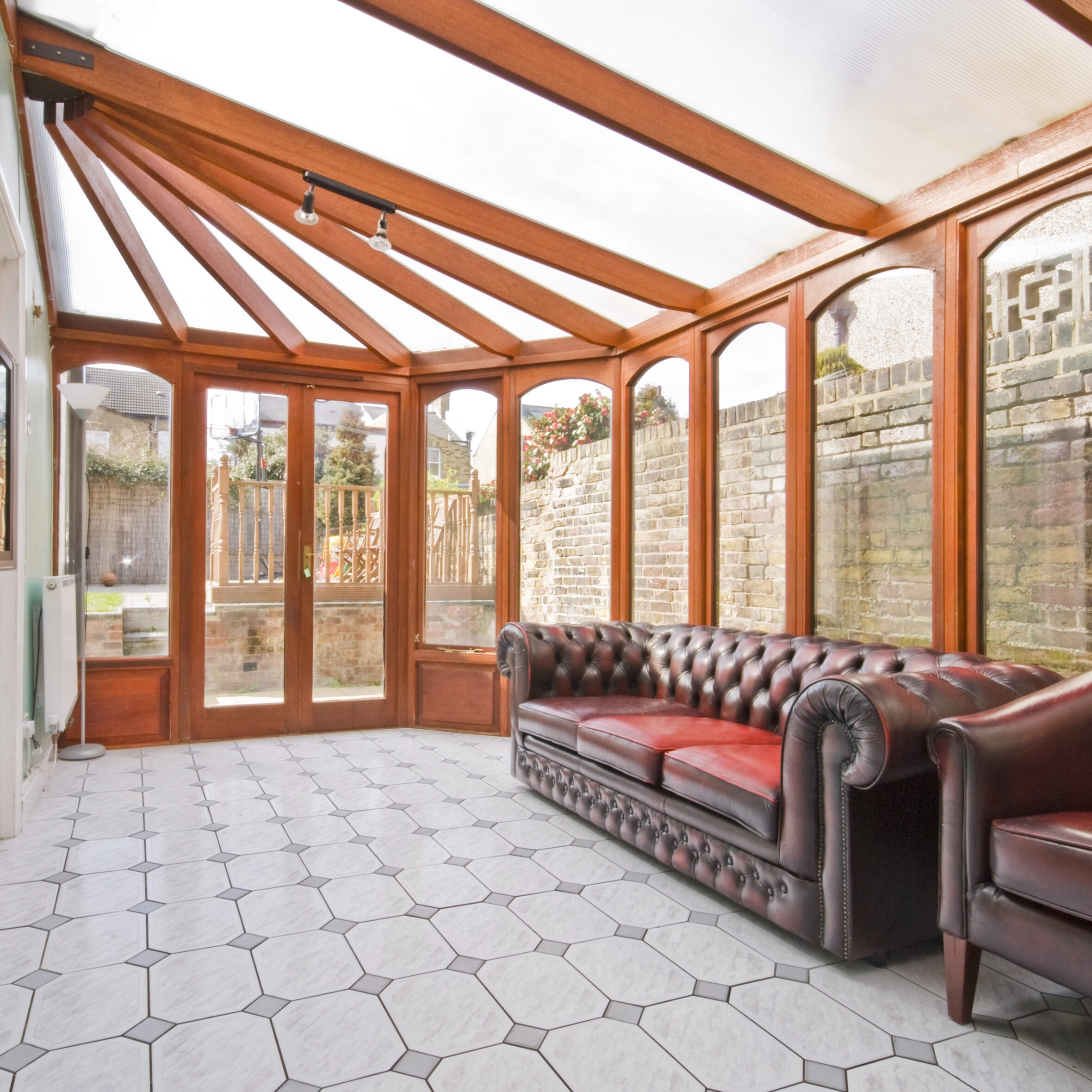 A sunlit room with large windows, leather sofa, and armchair. The room features a tiled floor and a brick wall outside.