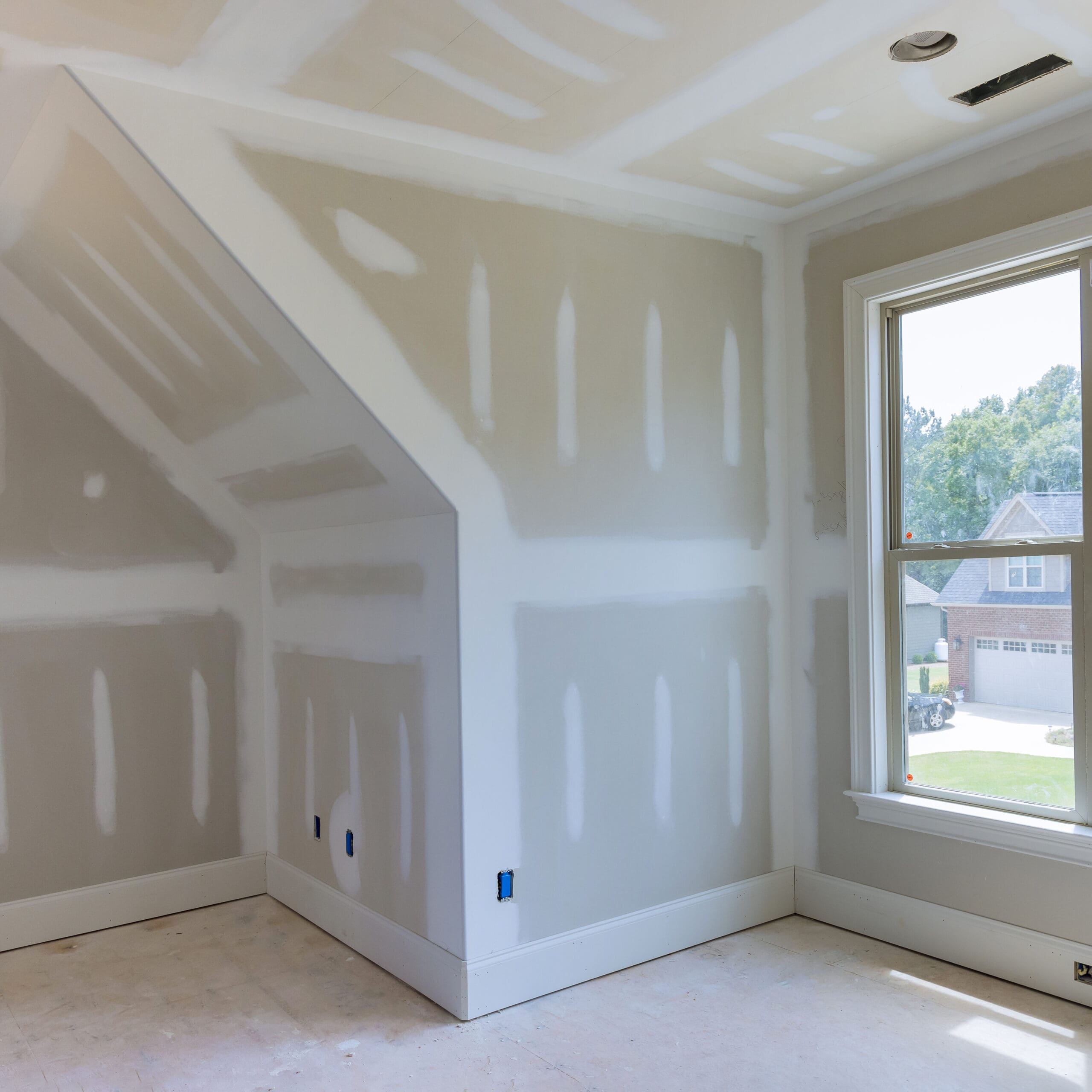 A room under construction with drywall installed and partially finished, featuring a large window overlooking a suburban neighborhood.