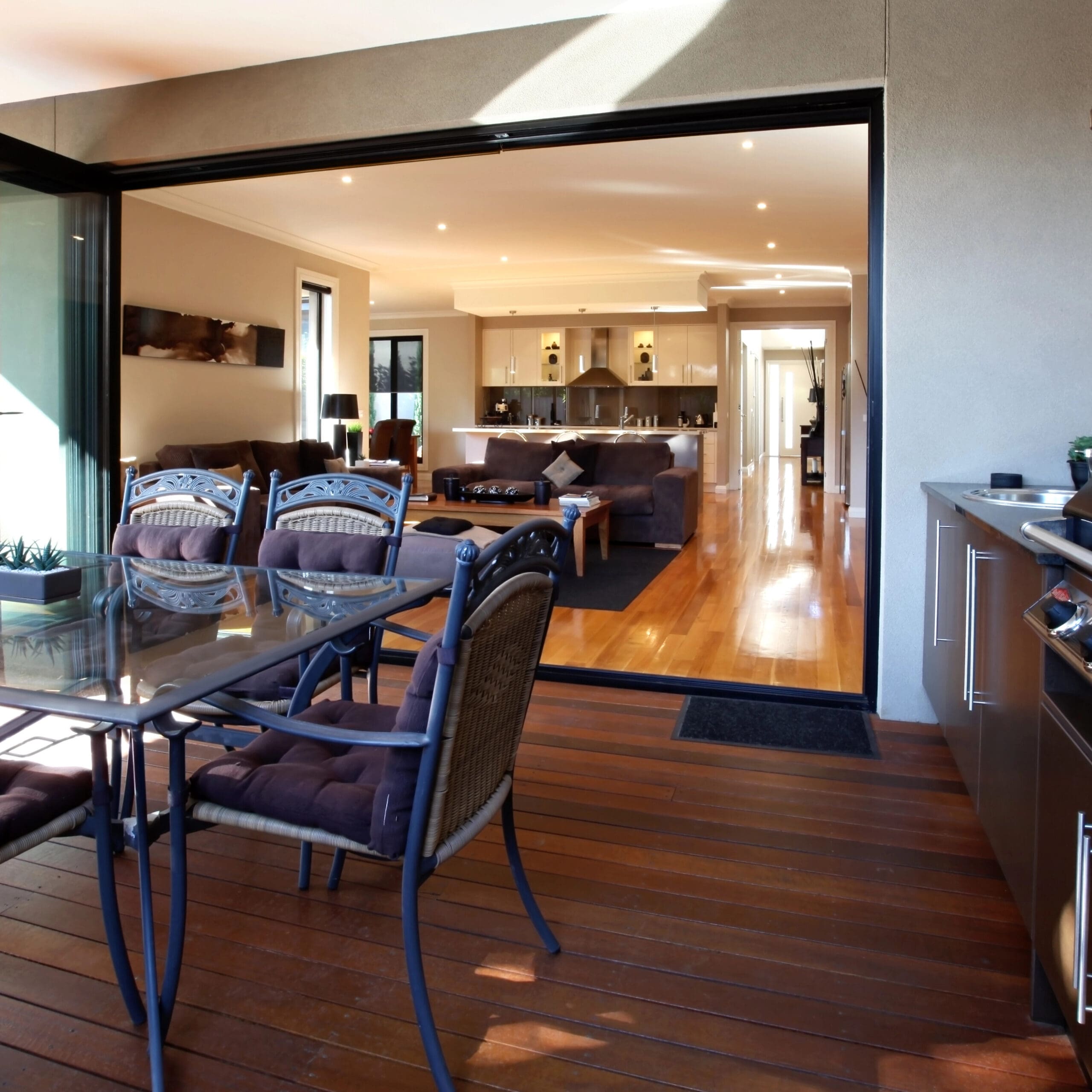 Modern open-plan living area with wooden flooring, encompassing a kitchen, dining space, and lounge area. The area extends to an outdoor patio featuring a dining table and barbecue grill.