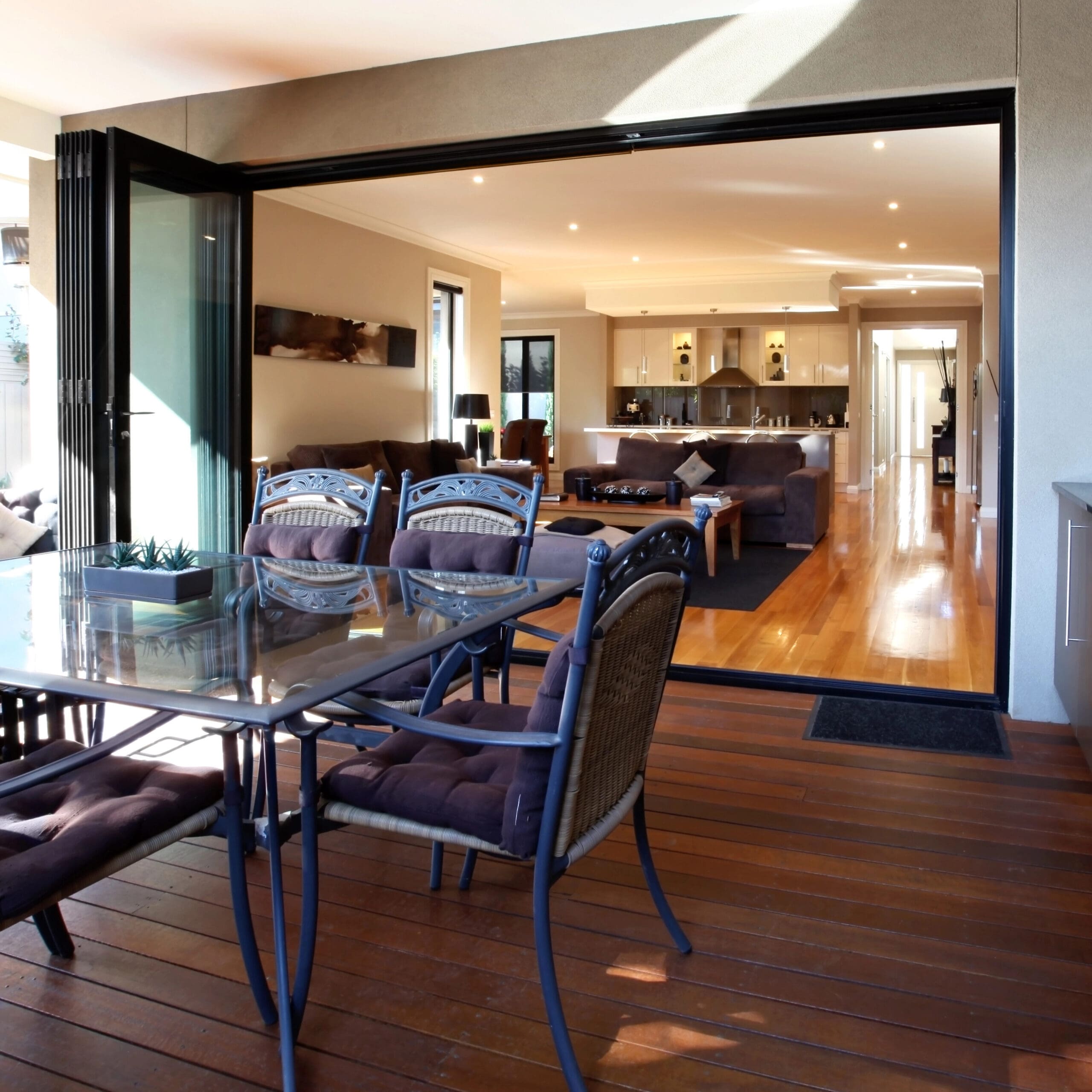 Modern open-plan living area with wooden flooring, encompassing a kitchen, dining space, and lounge area. The area extends to an outdoor patio featuring a dining table and barbecue grill.