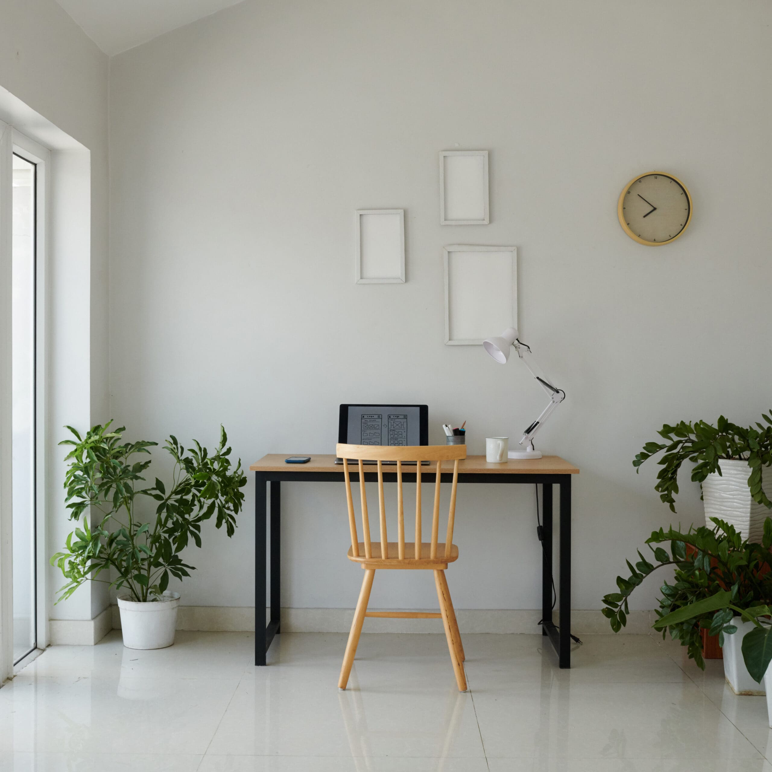 A minimalist workspace with a wooden desk, chair, laptop, desk lamp, and potted plants. Three empty frames and a clock hang on the white wall.