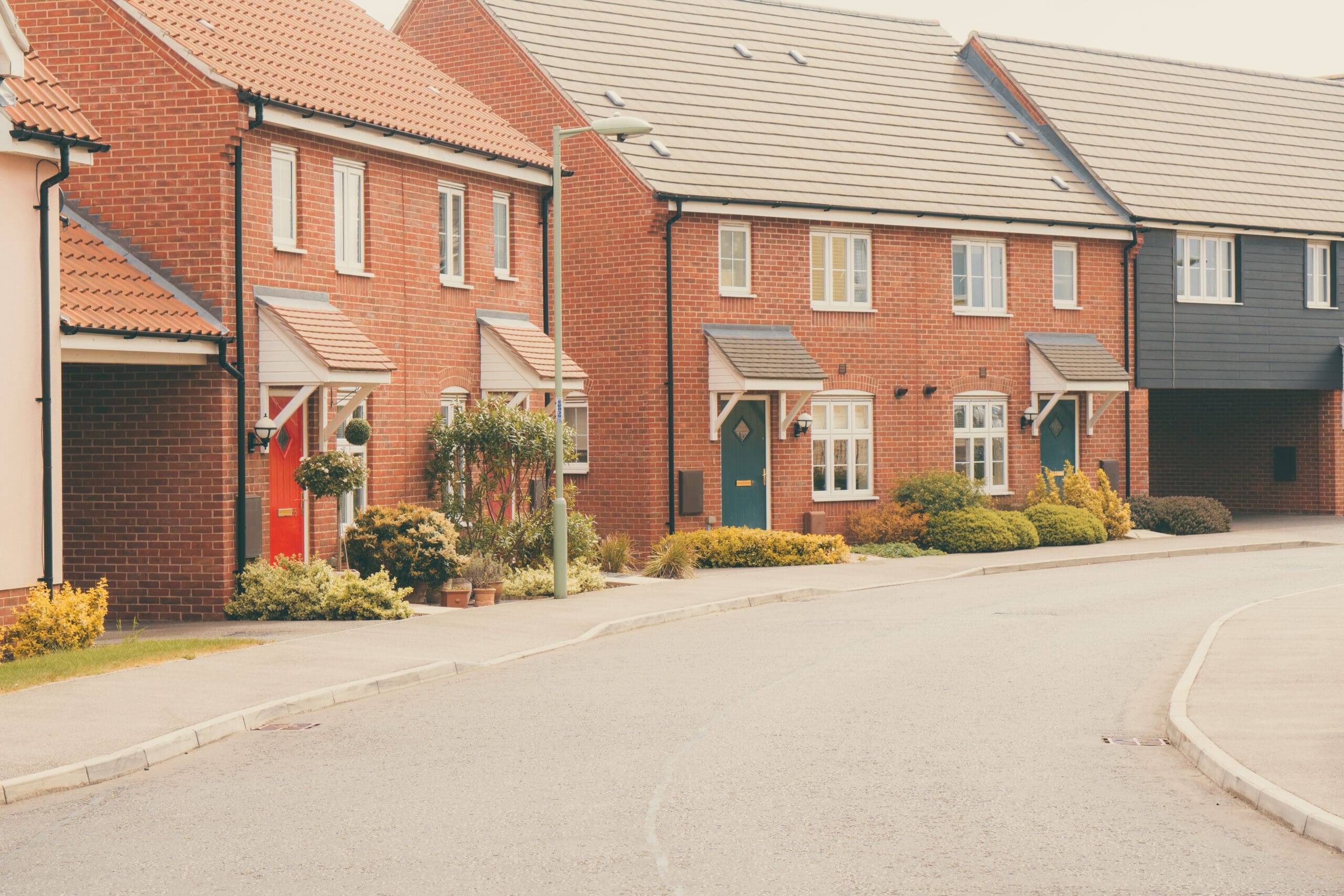 A row of modern red-brick townhouses with sloped roofs, small front gardens, and paved paths along a quiet, curved street.