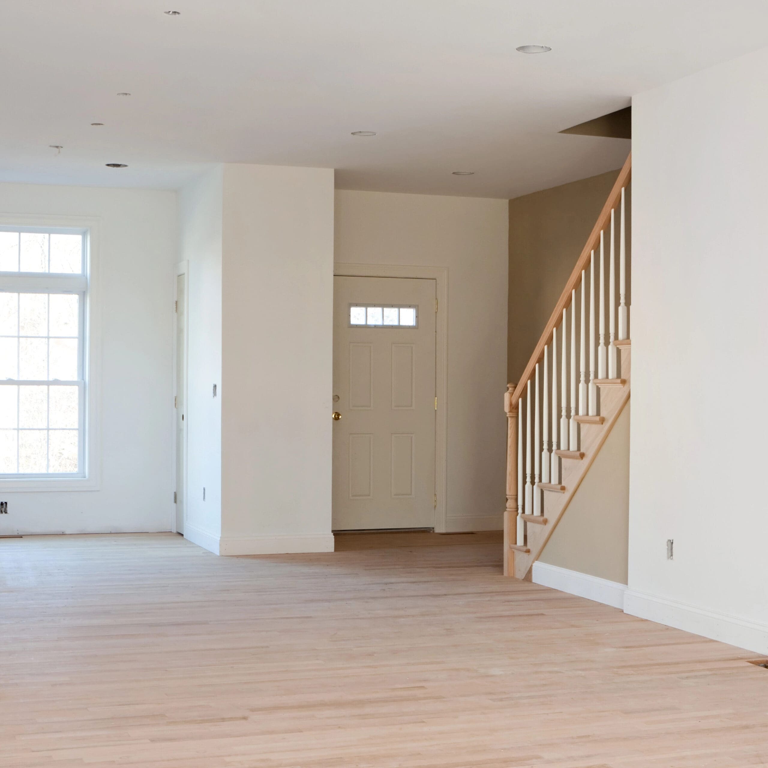 An empty room with light wooden floors, a window on the left, white walls, a staircase leading up on the right, and a closed beige door centrally located.