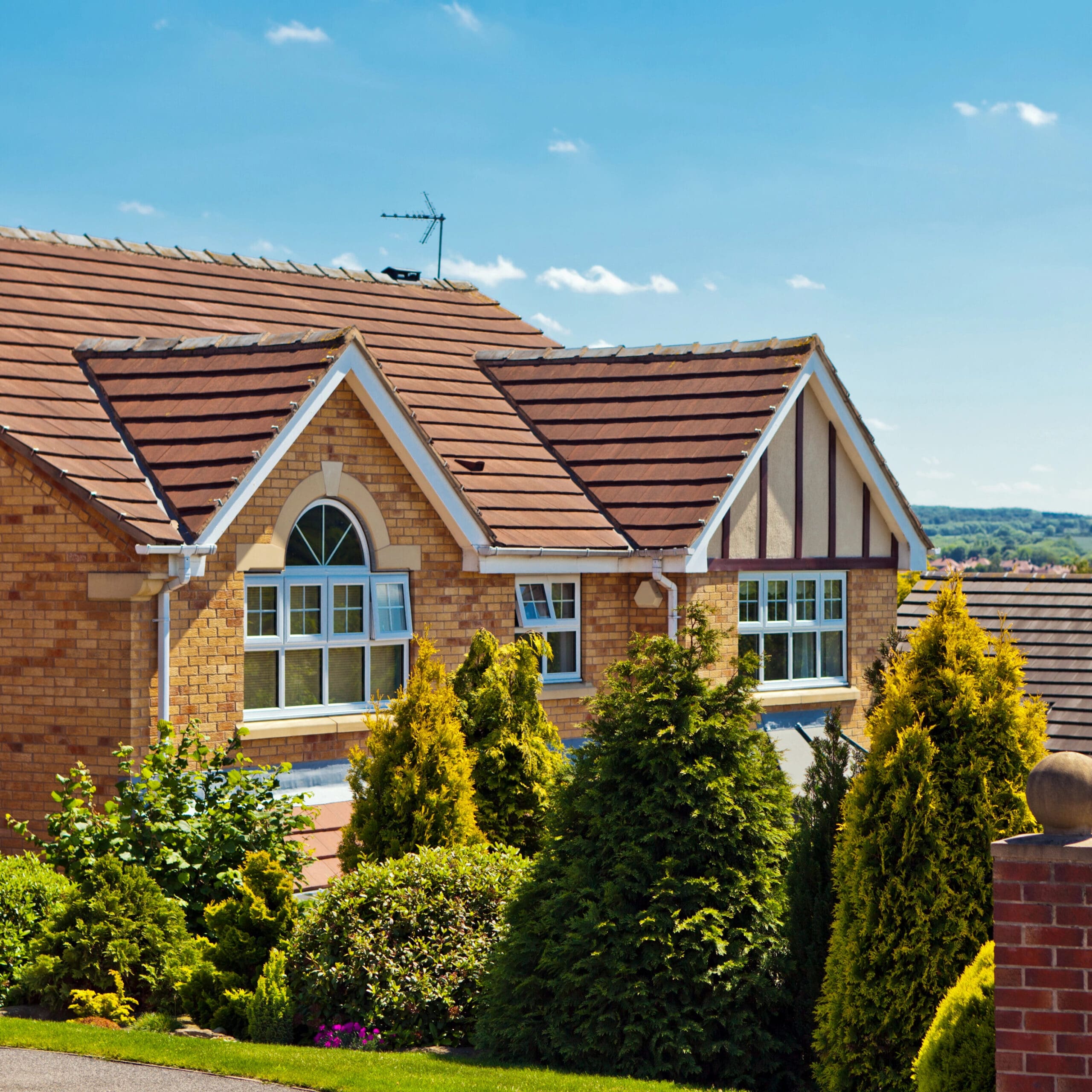 A brick house with a multi-gabled roof and decorative timber accents stands beside lush green shrubs and small trees under a clear blue sky.