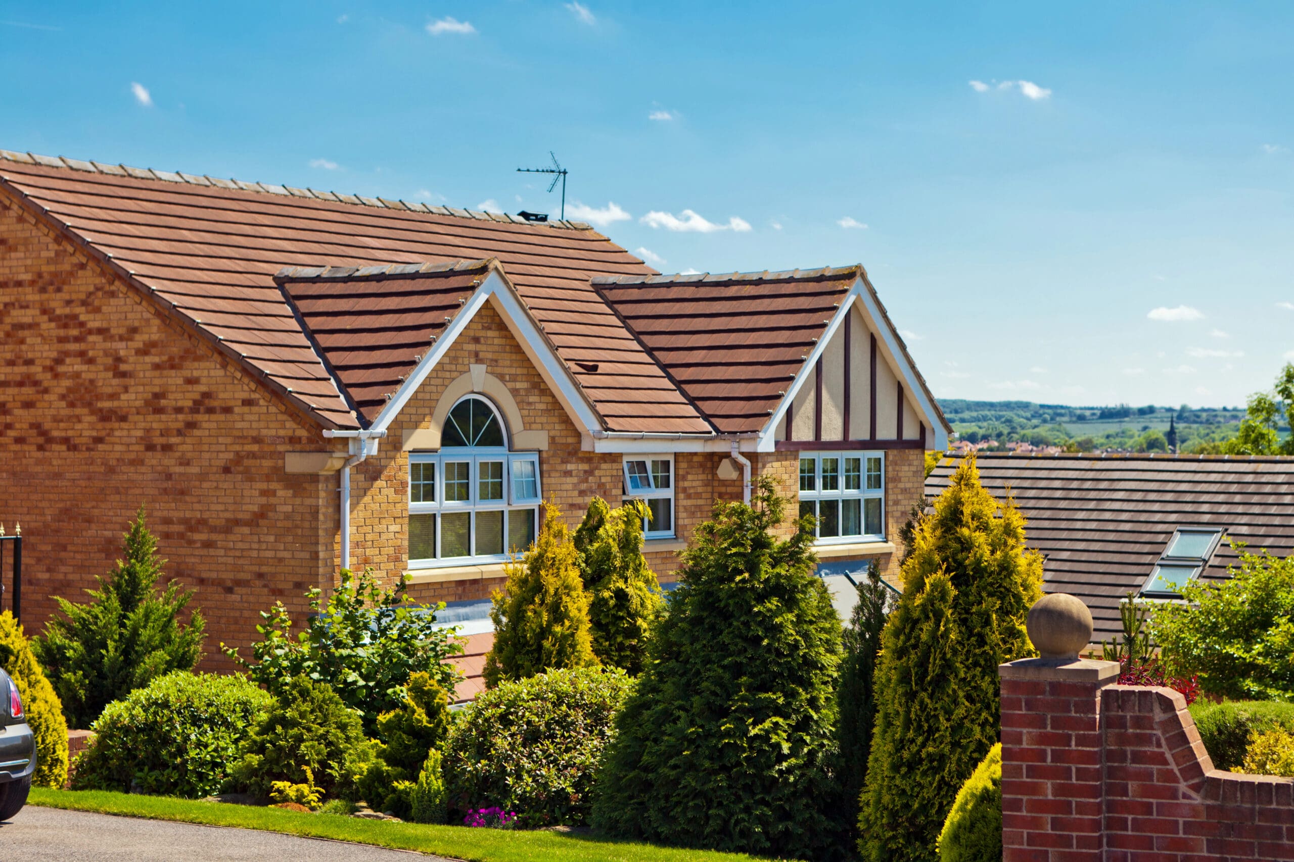 A two-story brick house with a gabled roof, surrounded by greenery and shrubs, under a clear blue sky.