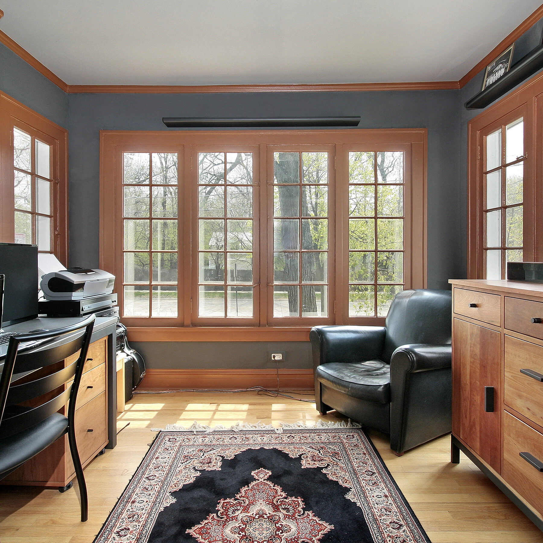 Home office with wooden furniture, a black chair, a desk with a printer, a shelf with model cars, and large windows letting in natural light.