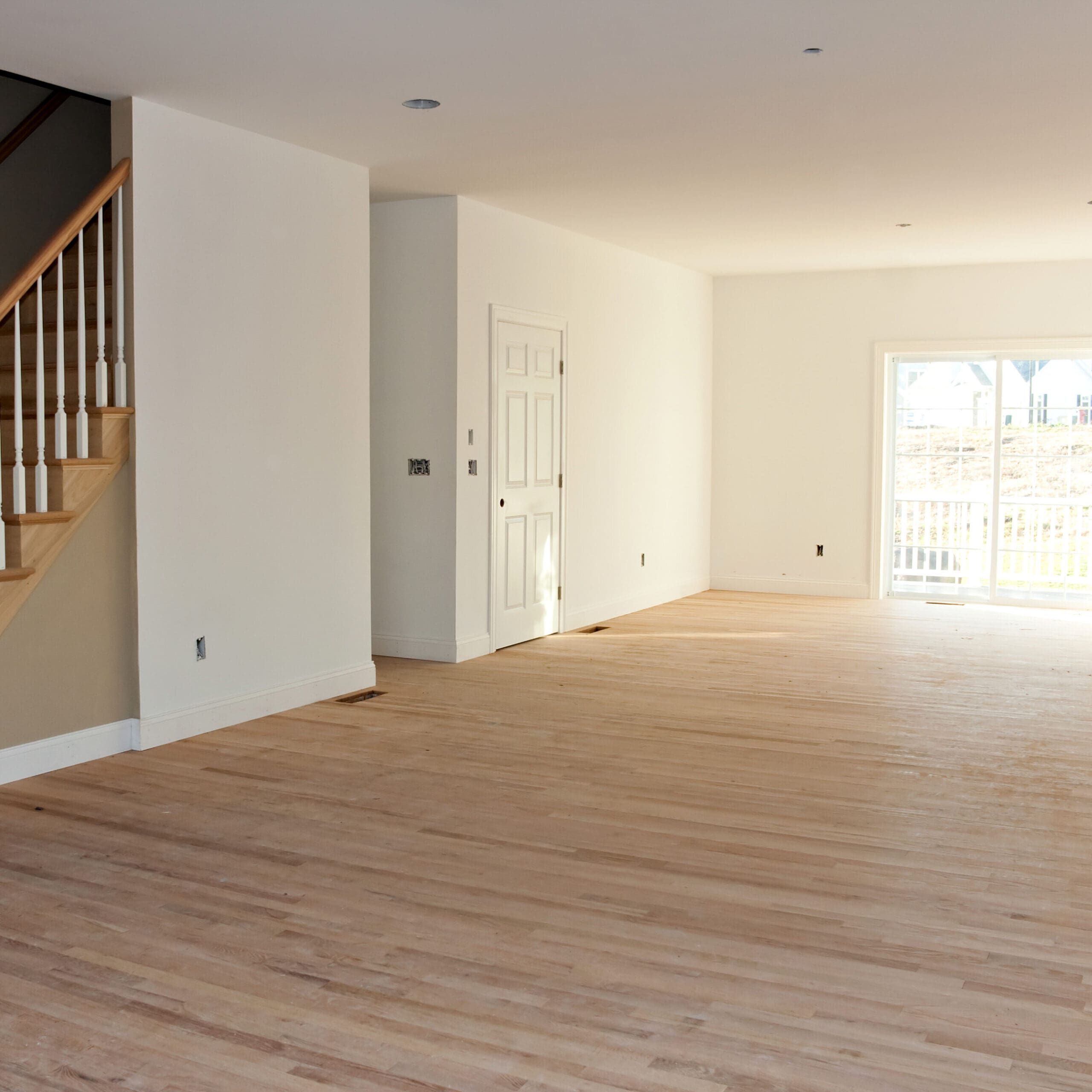 Empty room with freshly installed hardwood floors, a staircase with wooden railing on the left, white walls, and a door in the background leading to a bright outdoor area.