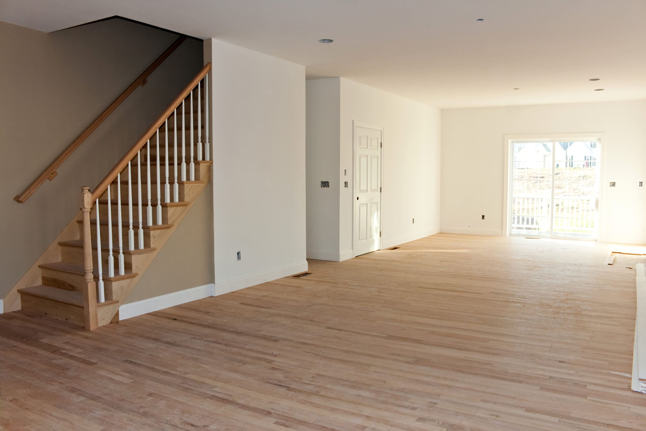 Empty room with freshly installed hardwood floors, a staircase with wooden railing on the left, white walls, and a door in the background leading to a bright outdoor area.