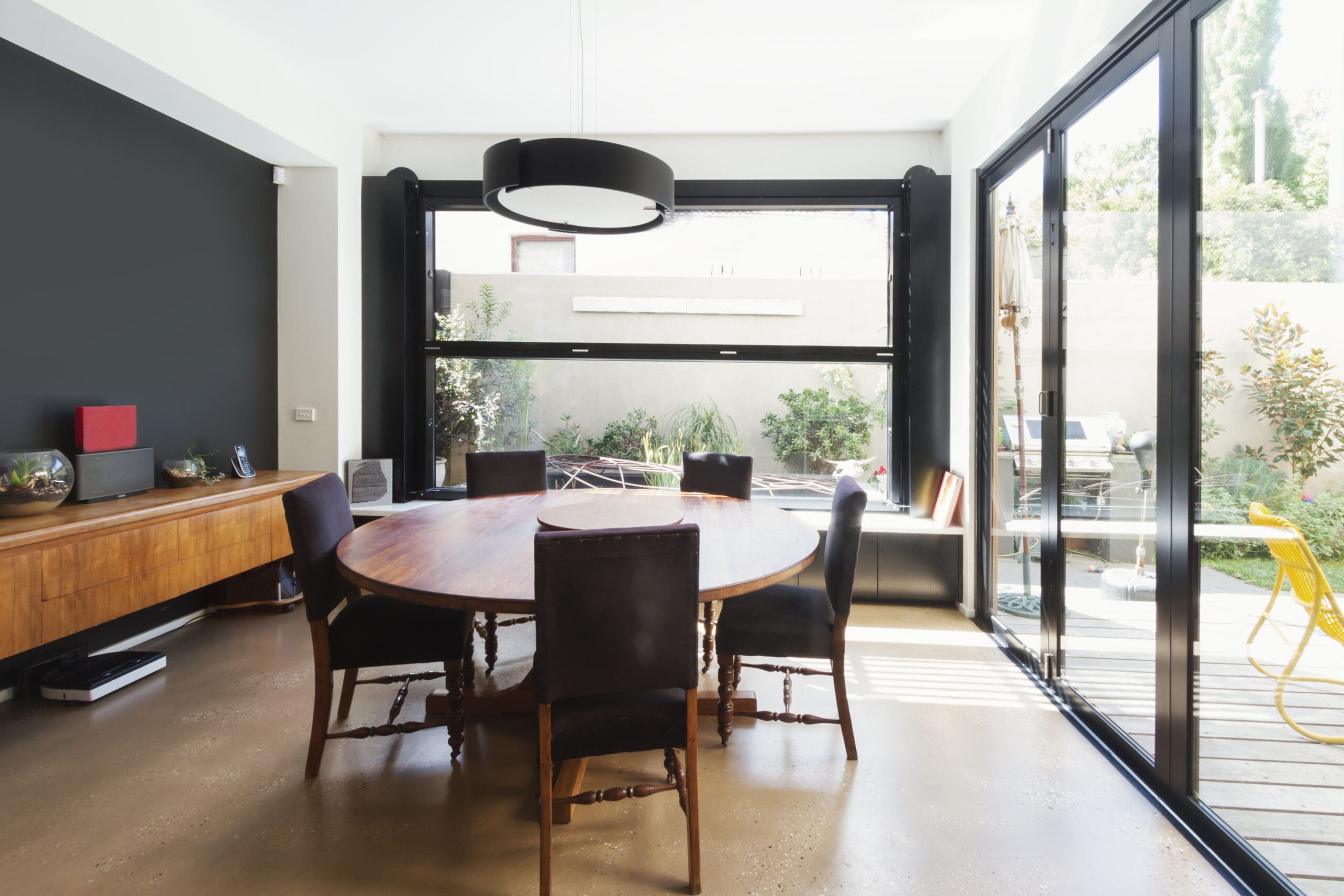 Modern dining room with a round wooden table, six chairs, large windows, black accent wall, and glass doors opening to a patio with plants.