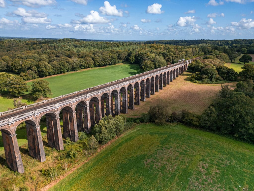 Arched brick railway viaduct crosses a green landscape with fields and trees under a partly cloudy sky.