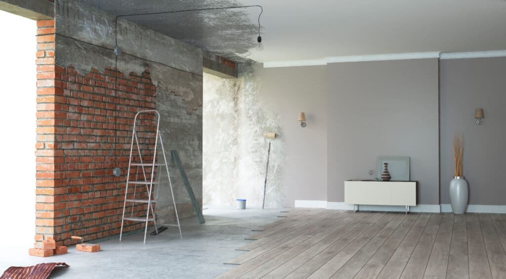 A room undergoing renovation with an unfinished brick wall, ladder, paint supplies, and partially installed wooden flooring.