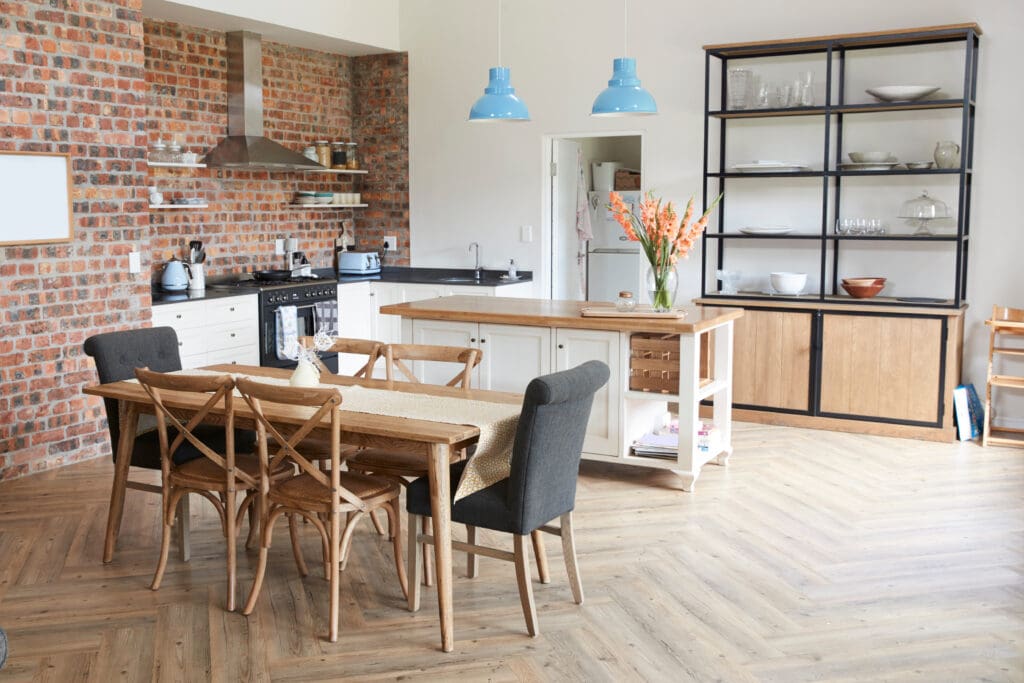 Modern kitchen and dining area with brick walls, wooden table and chairs, island with flowers, open shelving, and pendant lights.