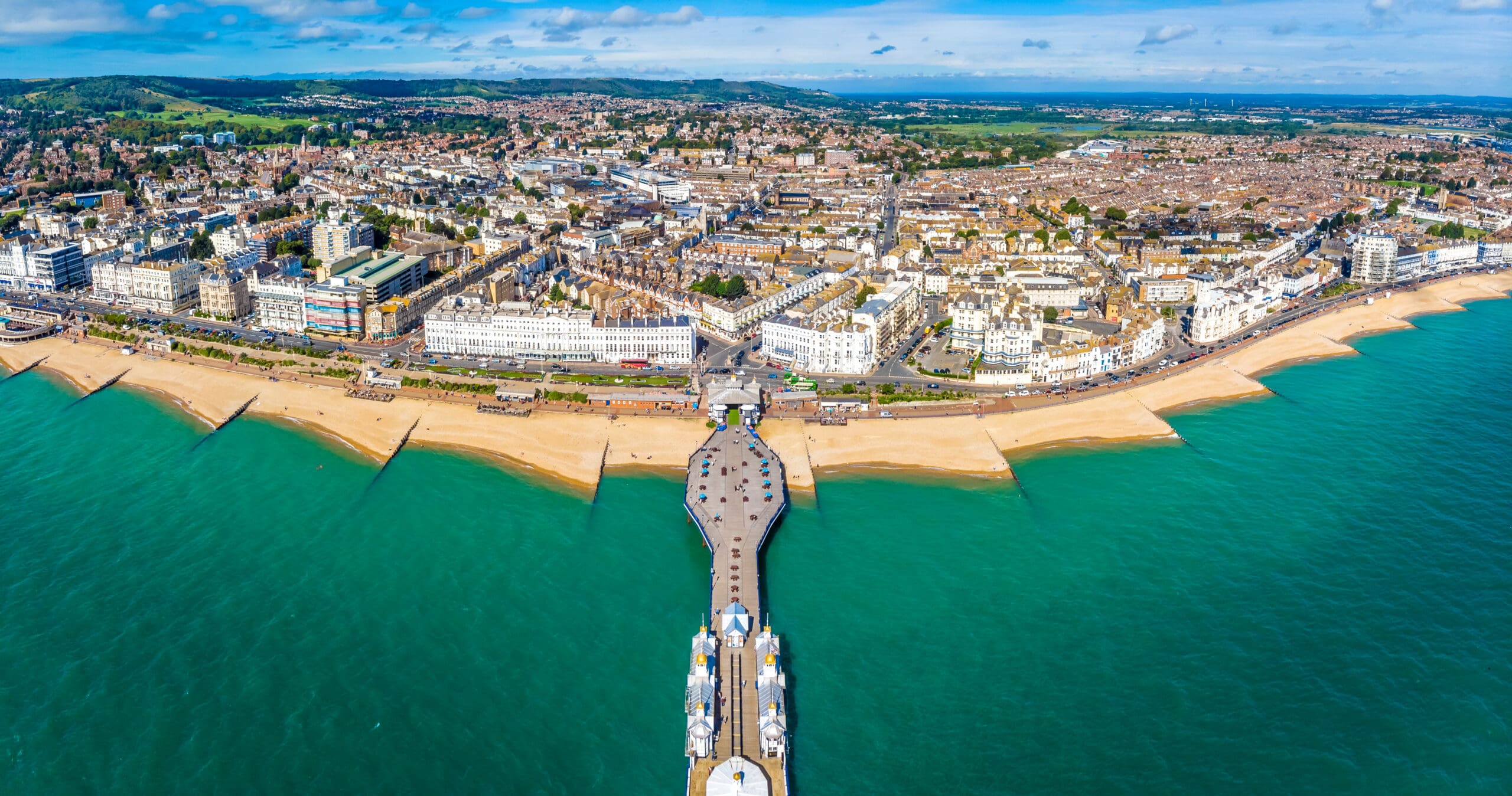 Aerial view of a seaside town with a long pier extending into the ocean, sandy beaches on both sides, and rows of buildings along the shoreline.
