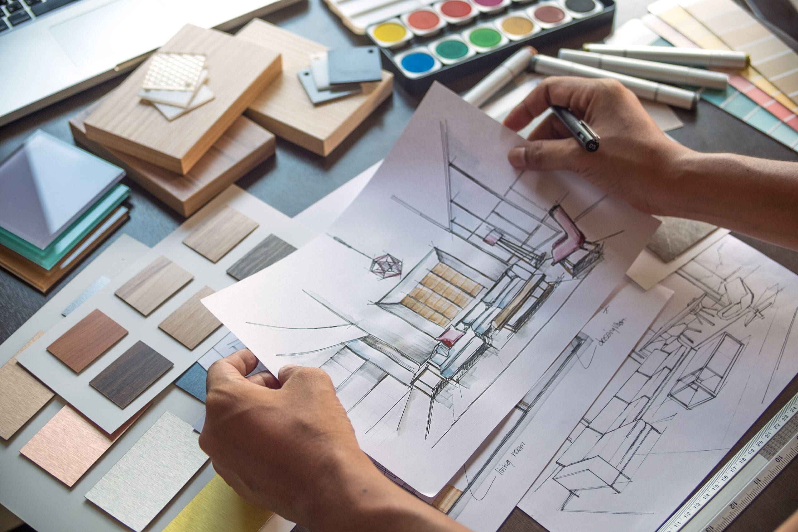 Person holding interior design sketches over a desk with material samples, color swatches, markers, and watercolor paints.