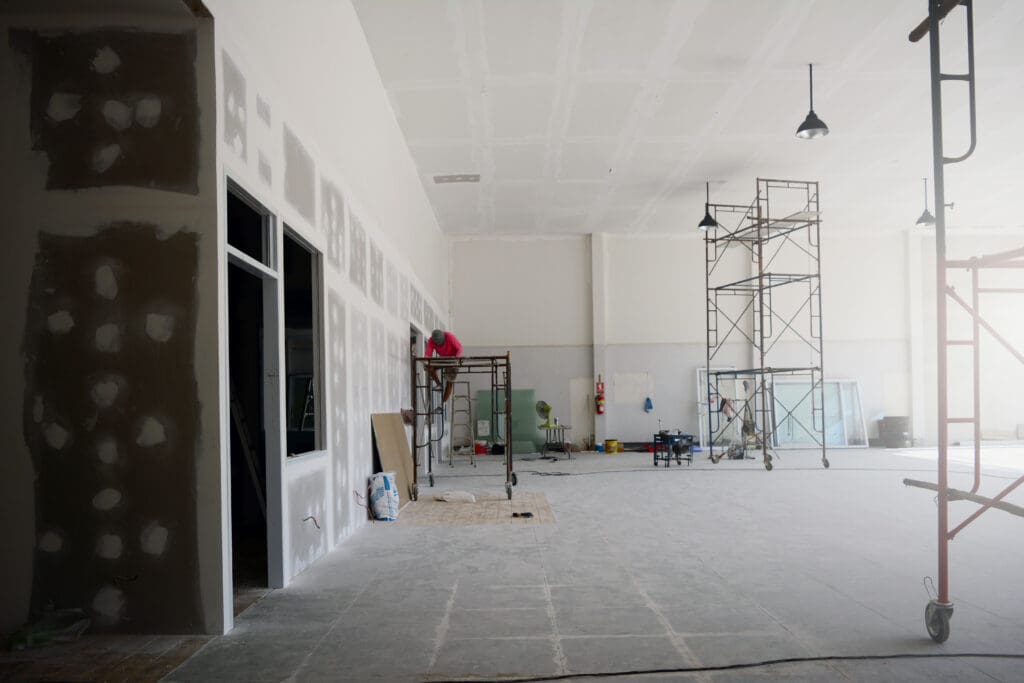 Large indoor space under construction with unfinished drywall, exposed scaffolding, construction materials, and workers present.