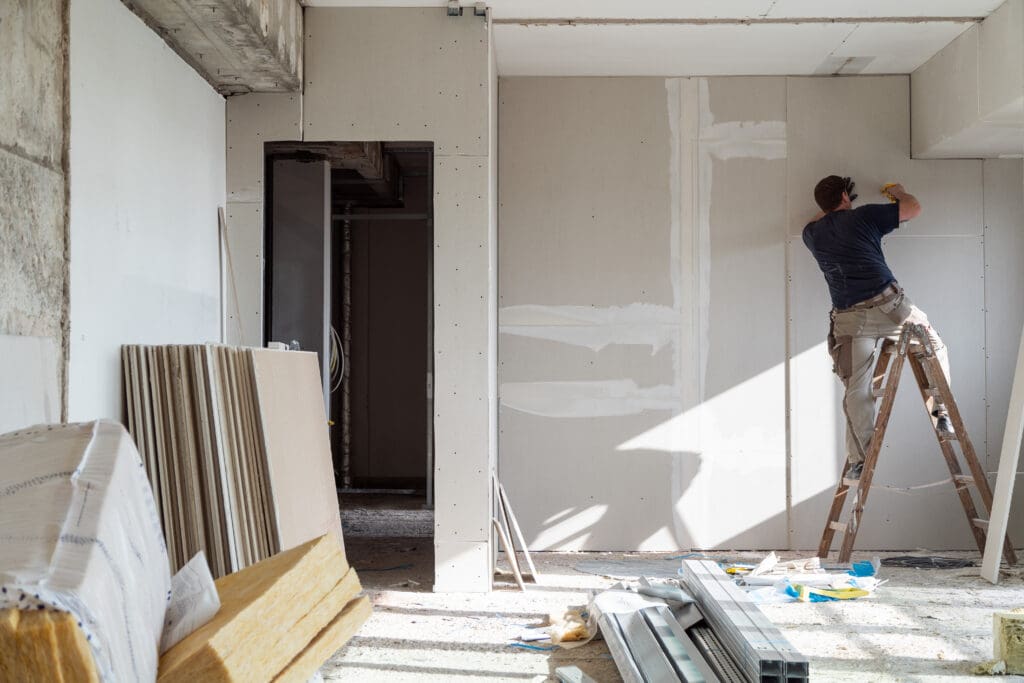 A person stands on a ladder installing or finishing drywall in a room under construction, with building materials stacked nearby.