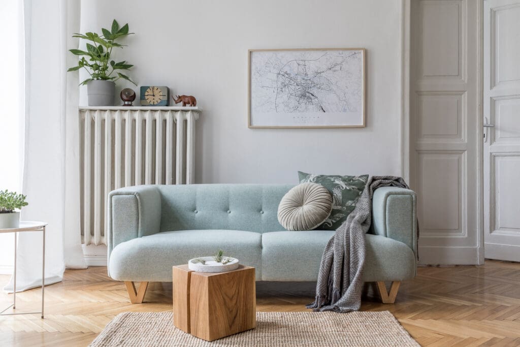 A light blue sofa with pillows and a gray throw sits in a living room with a framed map, radiator, plants, and wooden accents on a parquet floor.