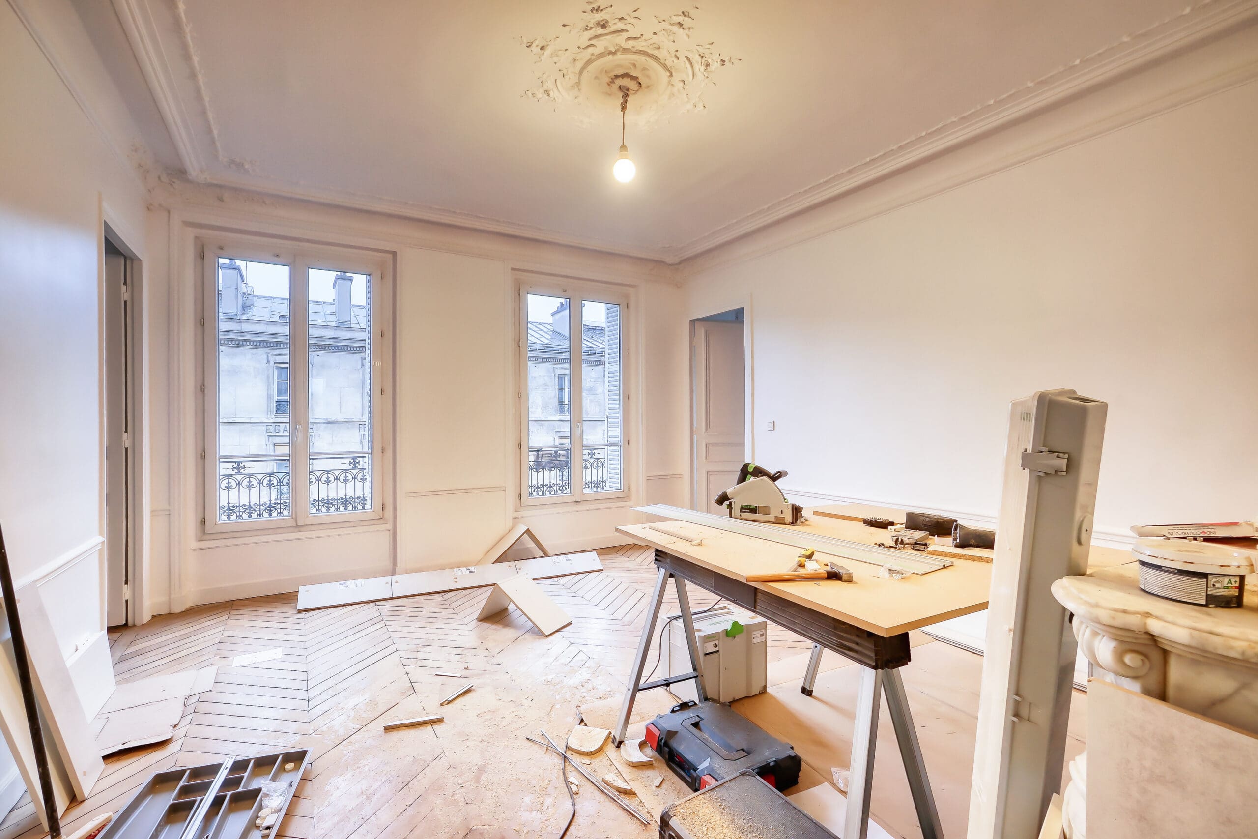 A partially renovated room with construction tools and materials, unfinished wooden flooring, and large windows letting in natural light.