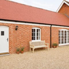 Red brick house with a white door, arched French windows, gravel driveway, wooden bench, and four potted plants in front.