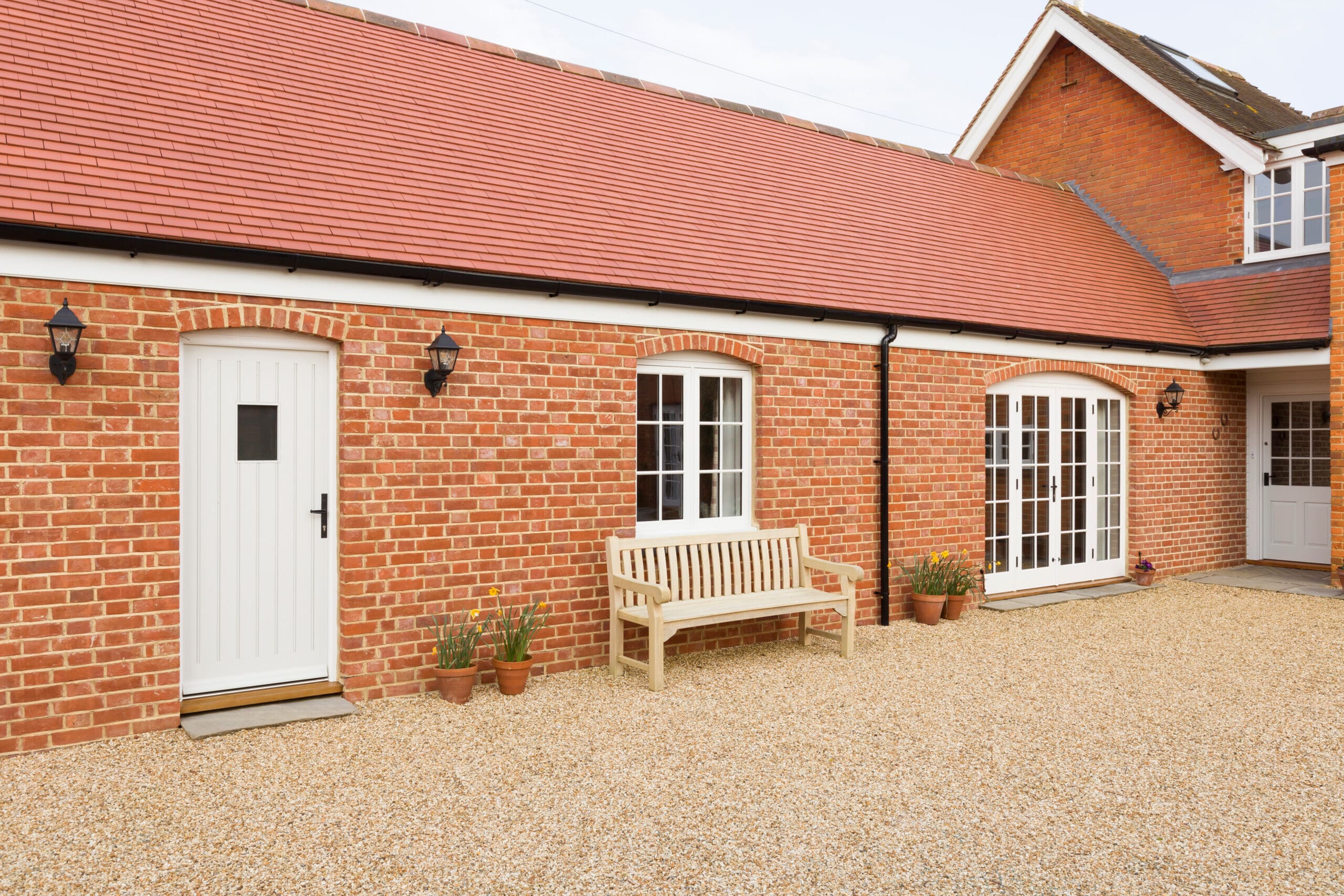 Red brick house with a white door, arched French windows, gravel driveway, wooden bench, and four potted plants in front.