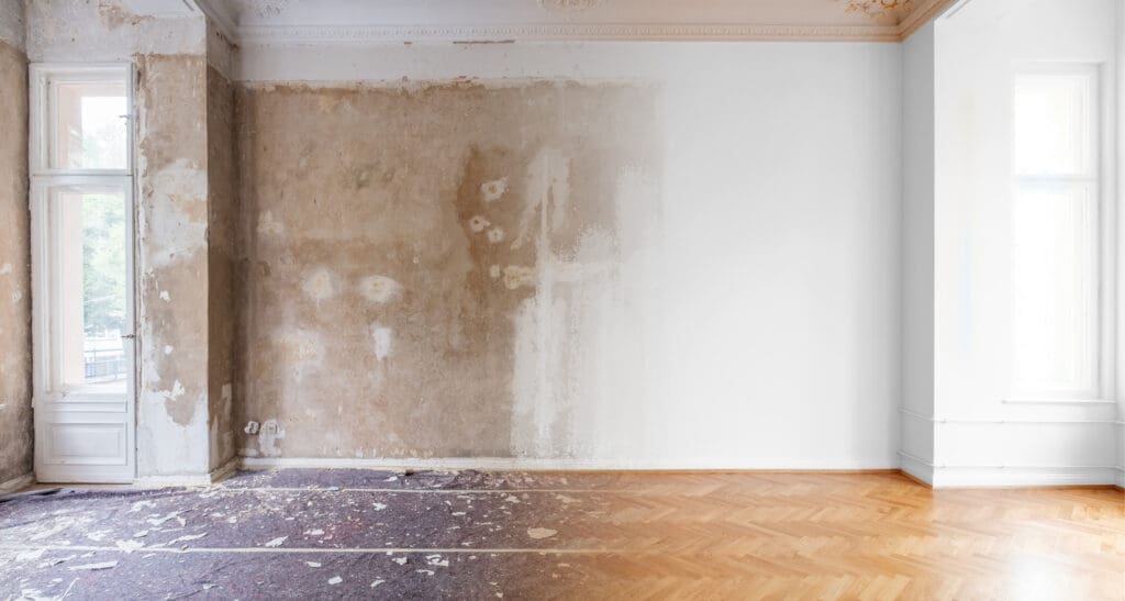 A partially renovated room with one wall stripped and patchy, old carpet on part of the floor, and new parquet flooring on the other side.