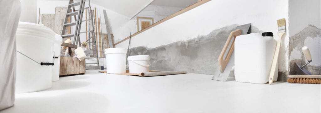 A room under renovation with white buckets, tools, a ladder, and paint supplies on the floor next to unfinished walls.