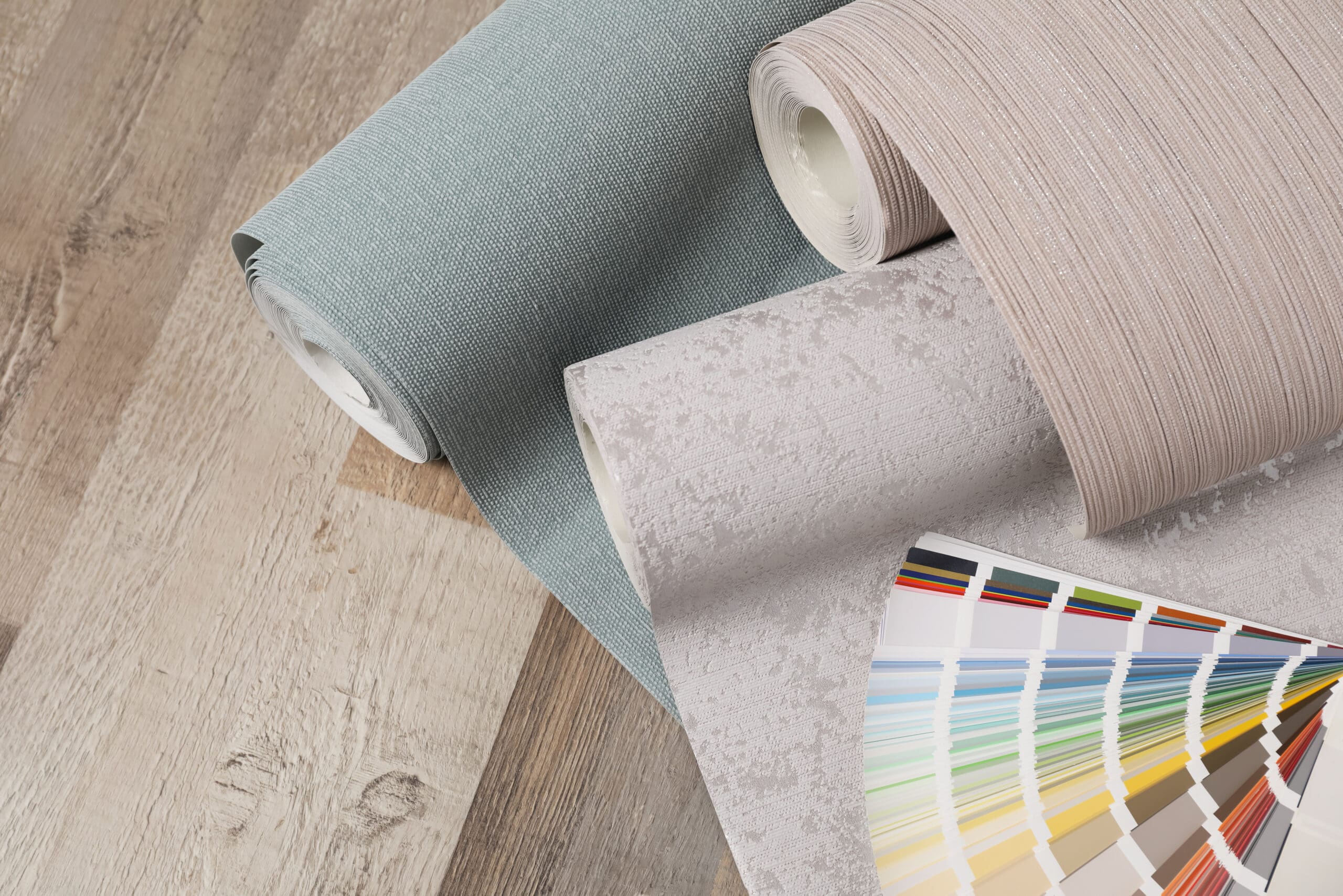 Three rolls of wallpaper in different textures and colors are placed next to a fan deck of paint swatches on a wooden surface.