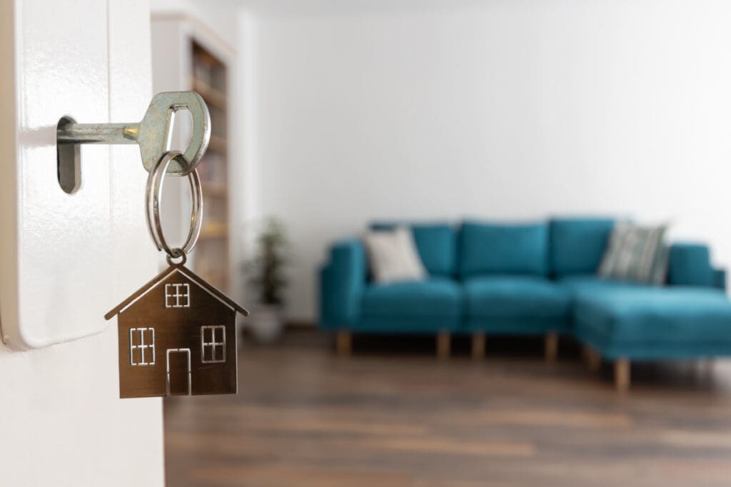 A key with a house-shaped keychain is inserted in a door lock; a blue sofa and blurred living room are visible in the background.