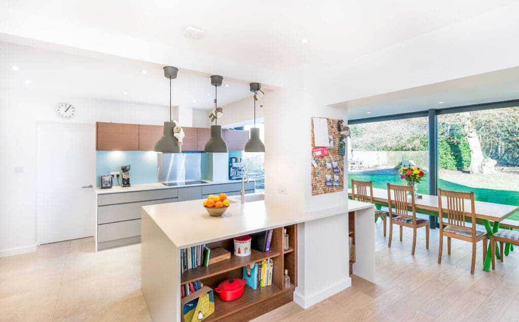 Modern kitchen with a central island, pendant lights, built-in bookshelf, dining table with chairs, large windows, and a view of a green backyard.