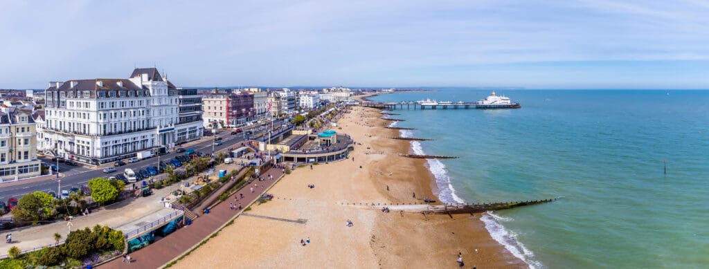 Aerial view of a seaside town with a pebble beach, a long pier extending into the sea, and rows of buildings lining the shoreline under a clear sky.