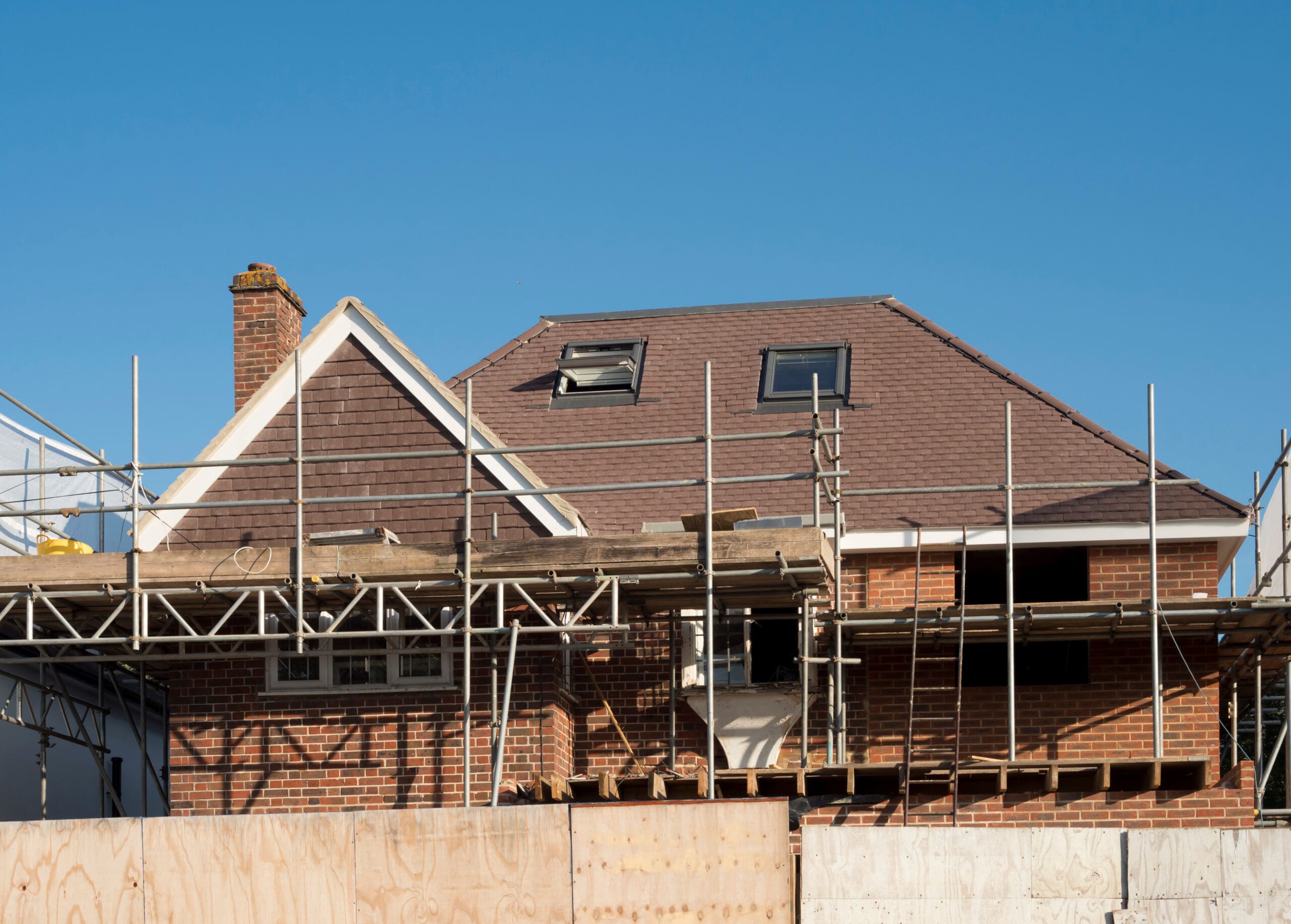 A brick house with a brown roof is under construction, surrounded by scaffolding and wooden barriers, set against a clear blue sky.