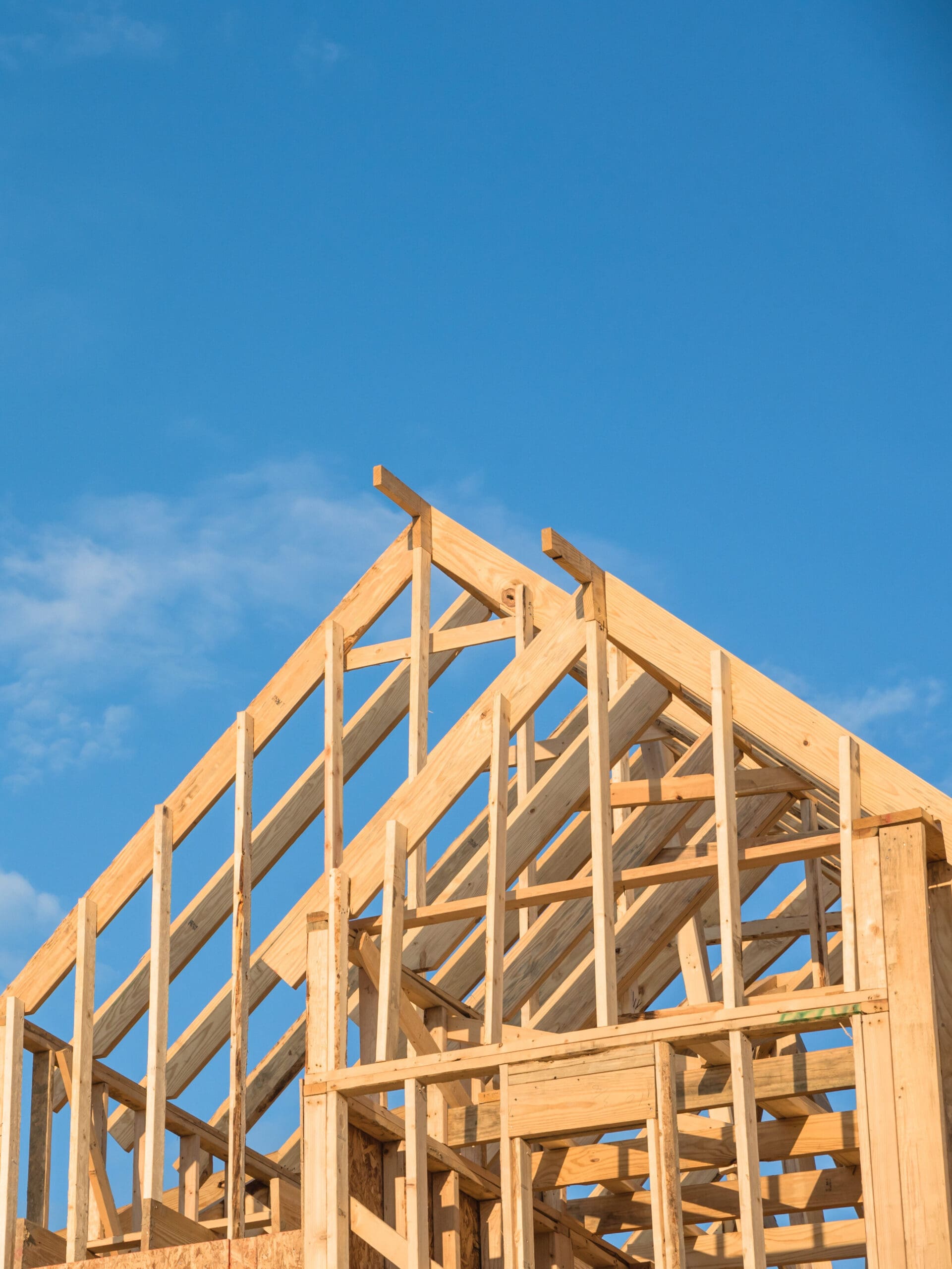 Wooden frame of a house under construction against a clear blue sky with some scattered clouds.