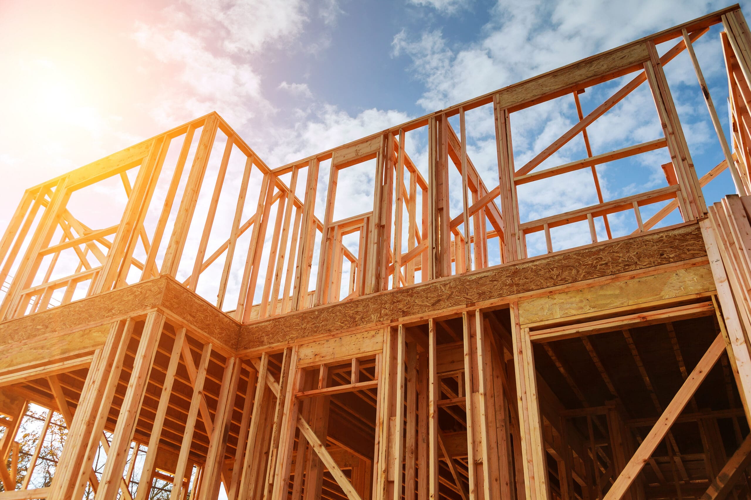 Wooden framing of a house under construction, with sunlight and blue sky in the background.